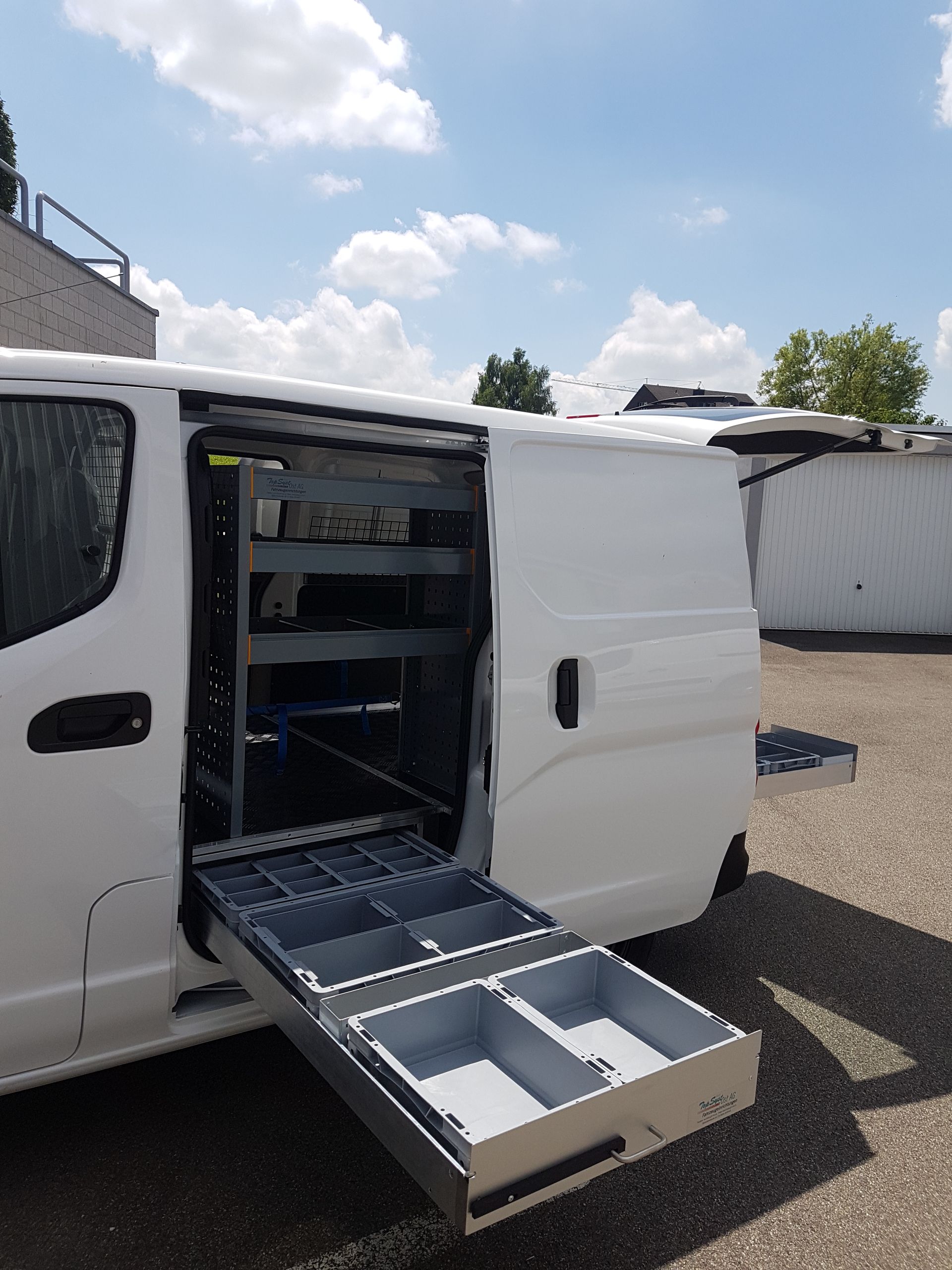 Weißer Lieferwagen mit offener Seitentür, hinter der sich ausziehbare Schubladen und Regale befinden, unter blauem Himmel.