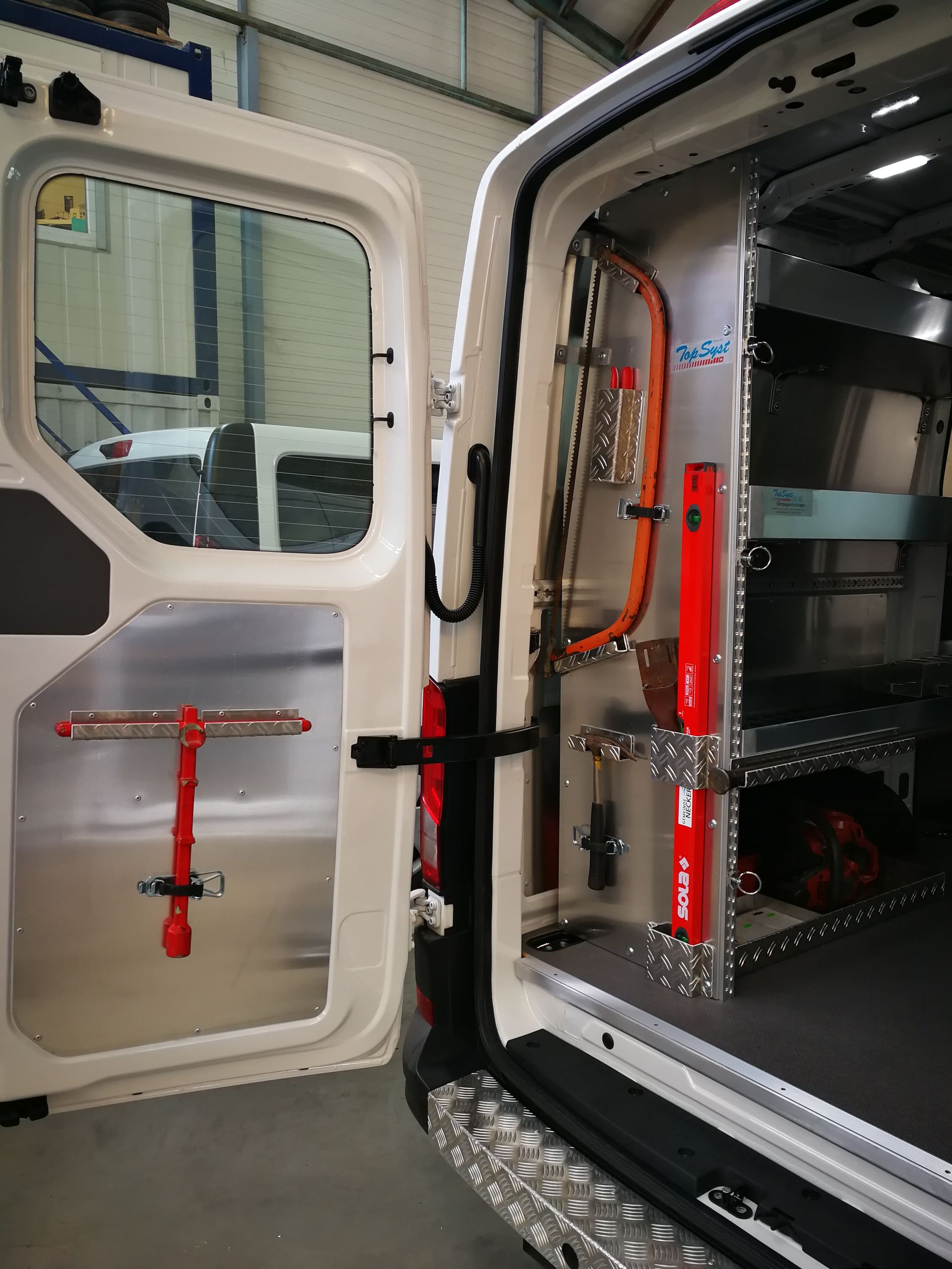 Die offene weiße Lieferwagentür gibt den Blick auf den Stauraum frei. Im Inneren sind Werkzeuge, Regale und rote Ausrüstung zu sehen.