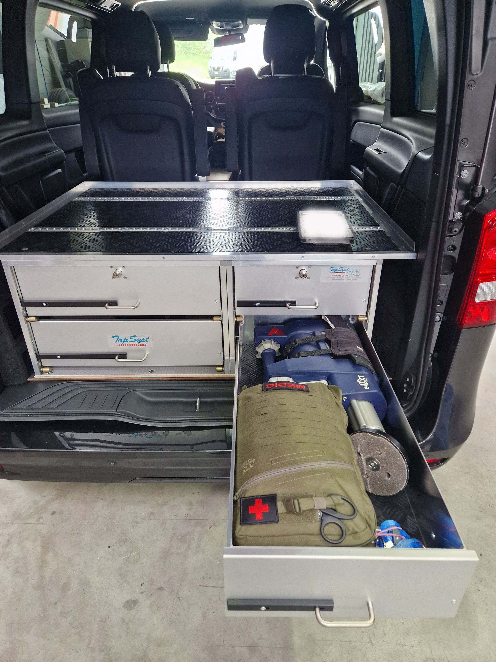 Innenraum eines schwarzen Lieferwagens mit offenen Schubladen, in denen Werkzeuge und ein Erste-Hilfe-Kasten zu sehen sind.