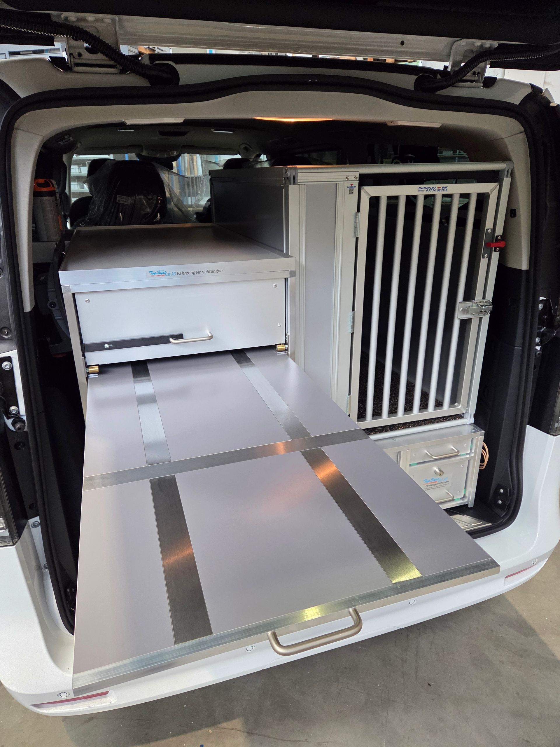 Ein Van-Innenraum mit Hundebox, Schubladen und ausziehbarer Plattform. Überwiegend in Silber und Weiß gehalten.