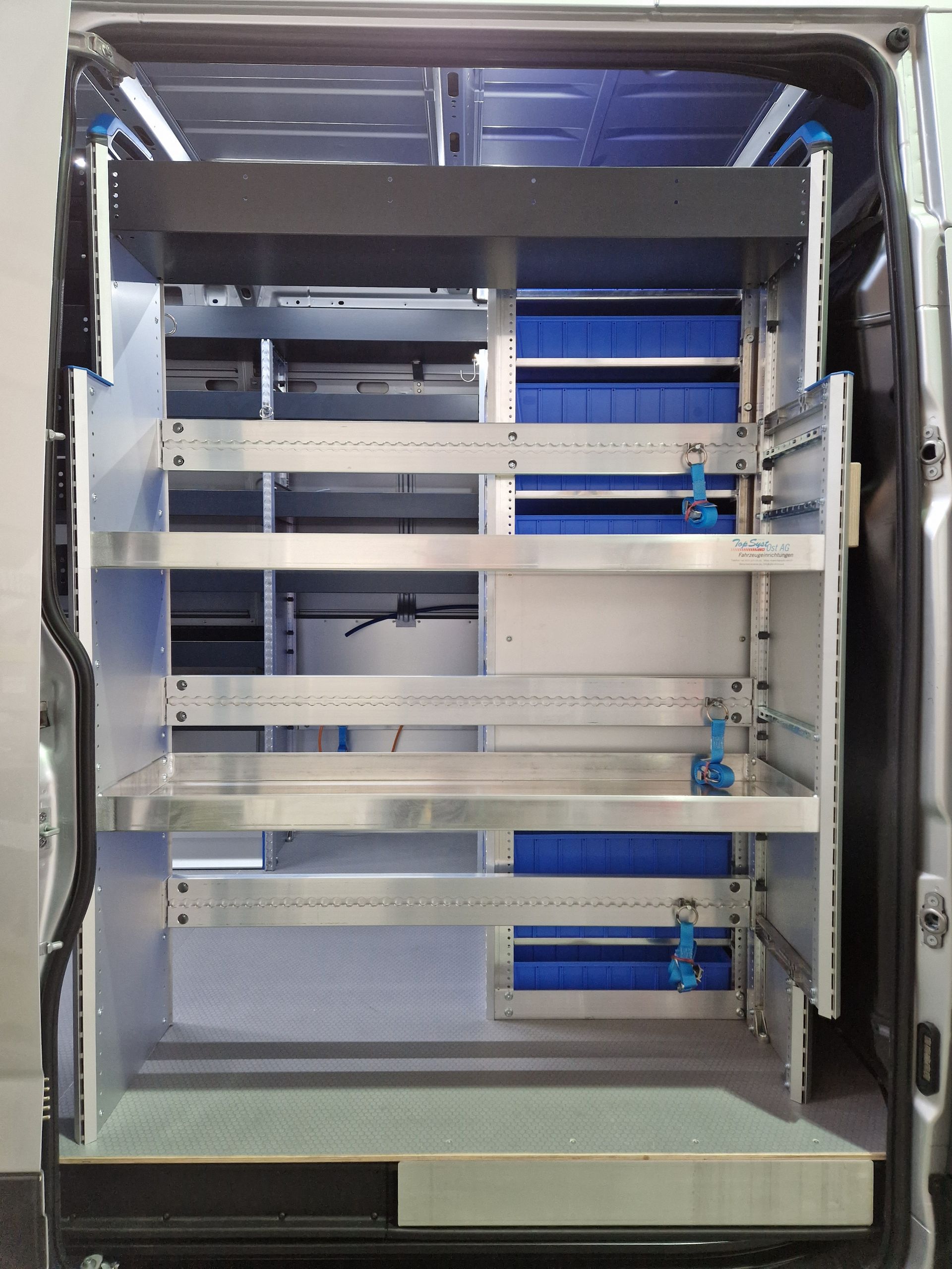 Innenraum eines Lieferwagens mit Regalen und Stauraum, überwiegend silberfarben mit blauen Behältern.