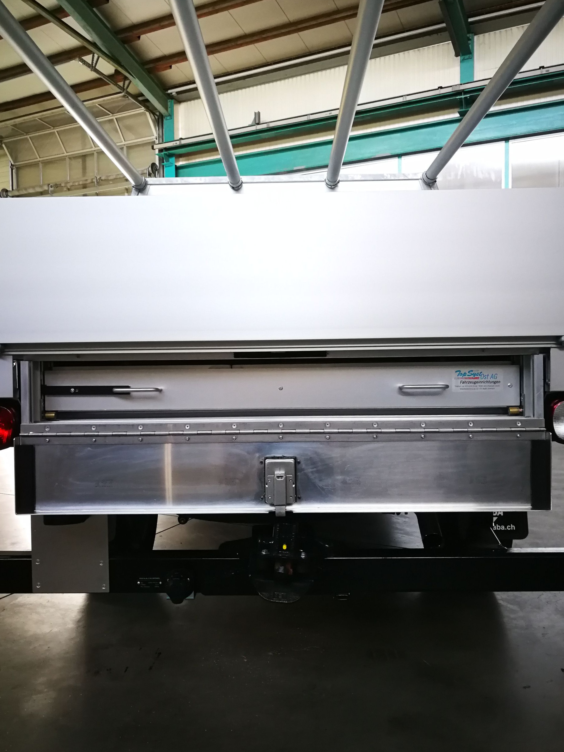 Rückseite eines weißen Lastwagens mit offenem Fach, Metallstoßstange und Dachträger.