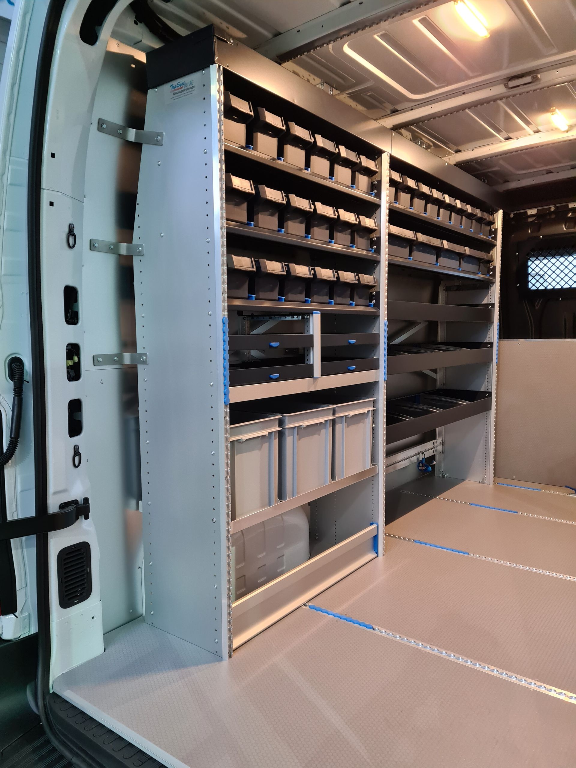 Innenansicht eines Lieferwagens mit Regalen. Die Regale enthalten Behälter und Vorräte. Grauer Boden und weiße Wände.
