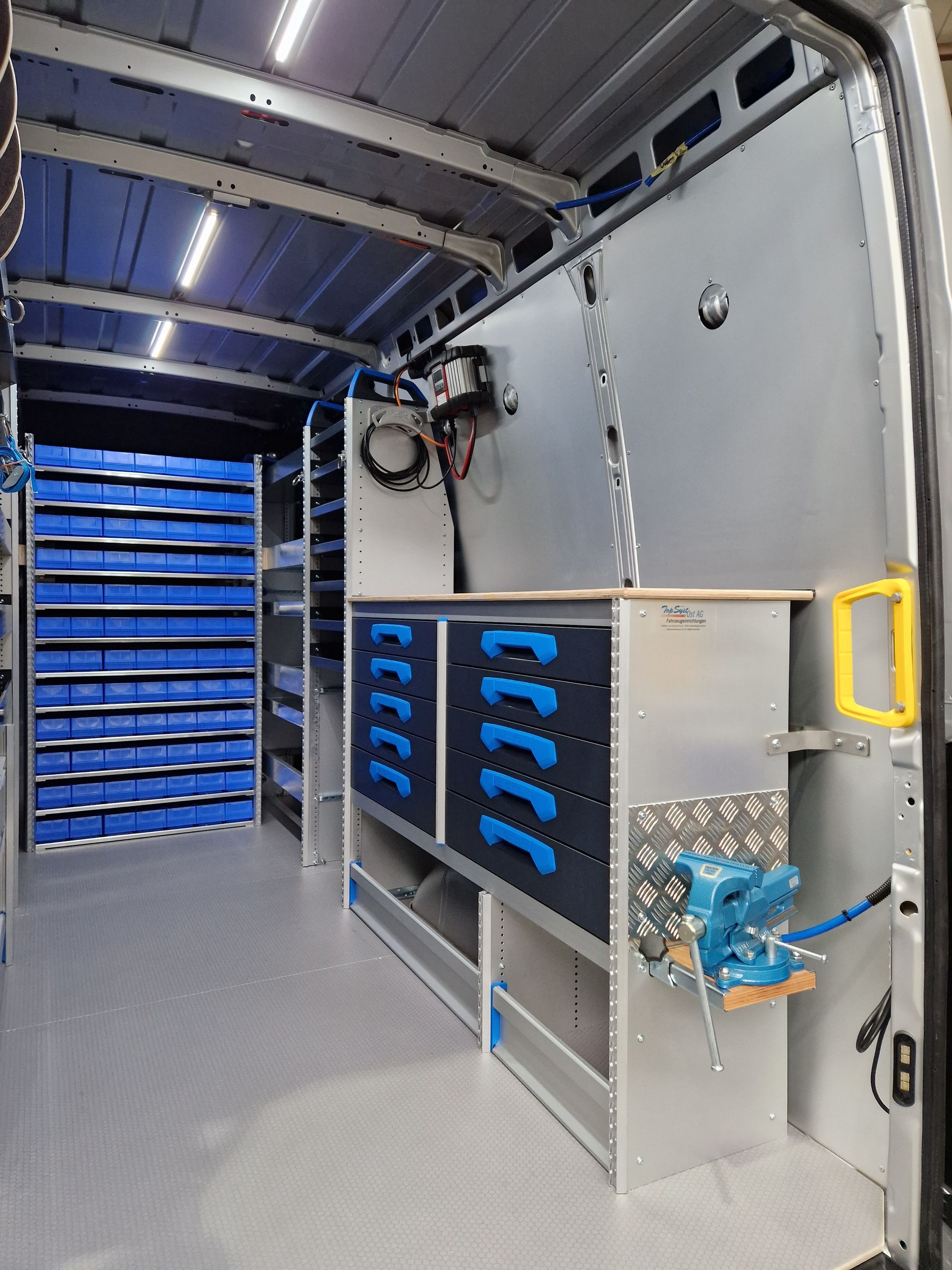 Innenansicht eines Lieferwagens, ausgestattet mit blauen Lagerbehältern, Schubladen und einem montierten Schraubstock.