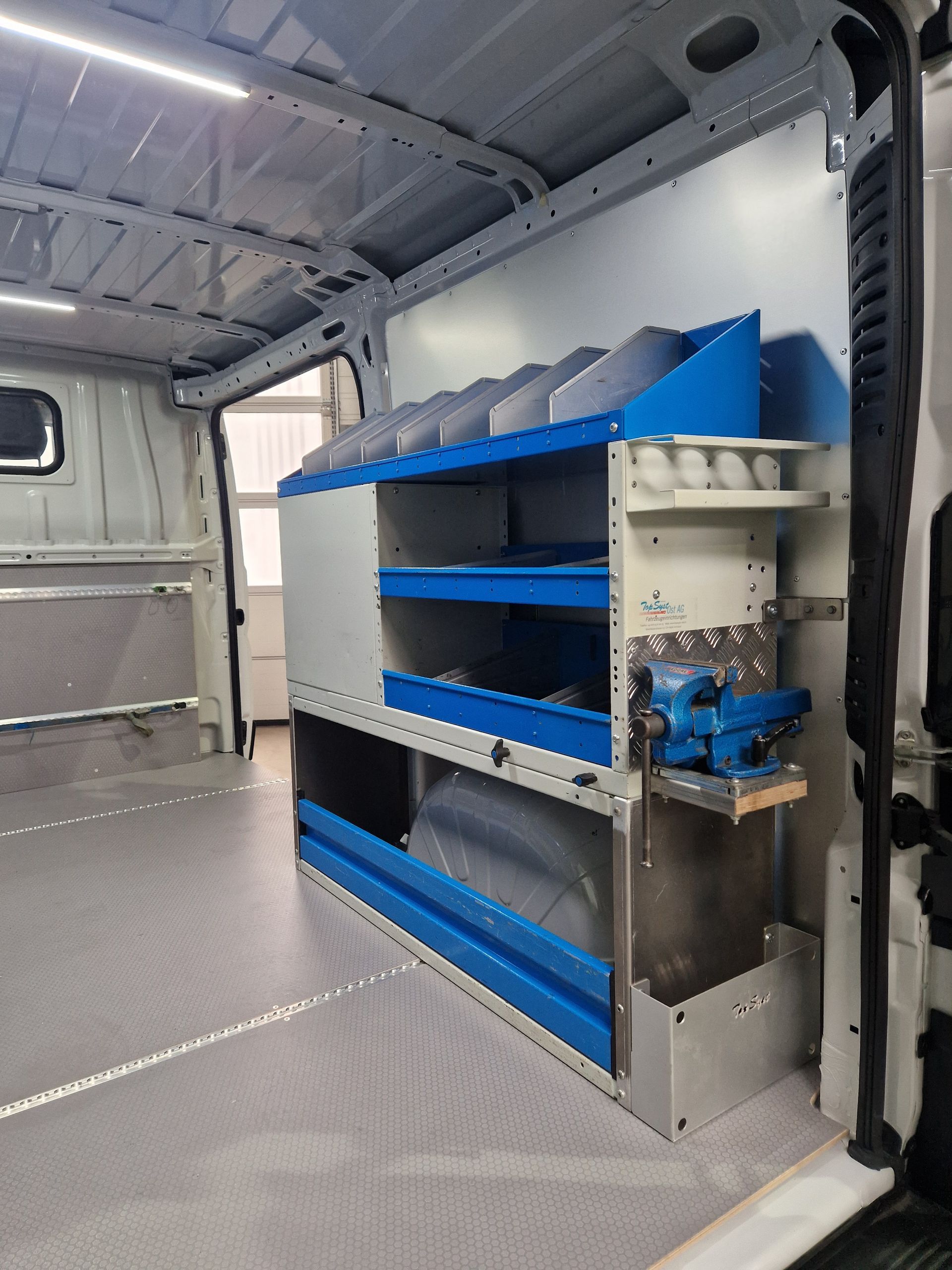 Innenraum eines weißen Lieferwagens mit Metallregal, blauen Behältern und Regalen, einem montierten Schraubstock.