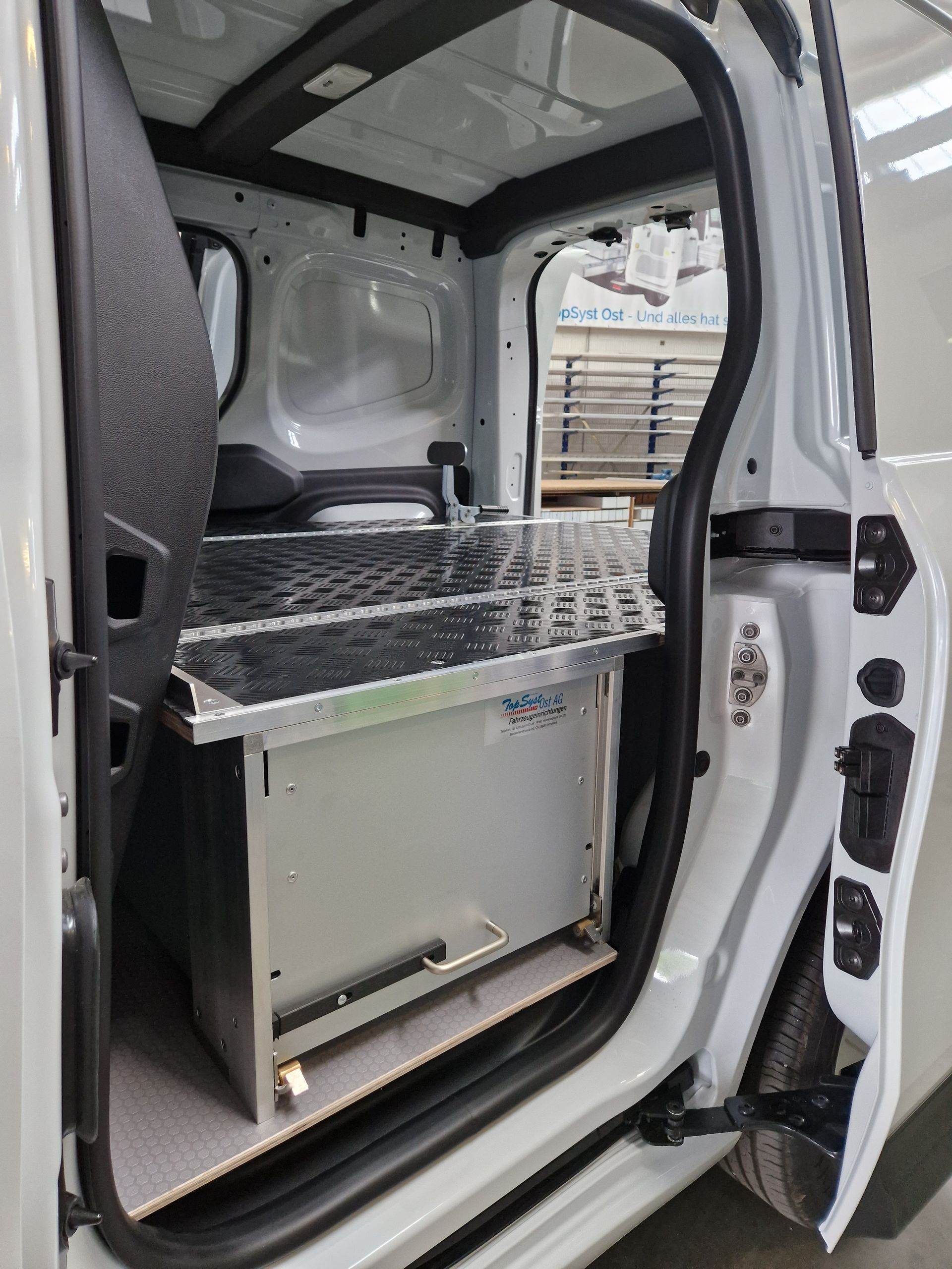Offener weißer Van-Innenraum mit eingebauter Schubladeneinheit aus Metall und Spüle, sichtbar durch die offene Tür.