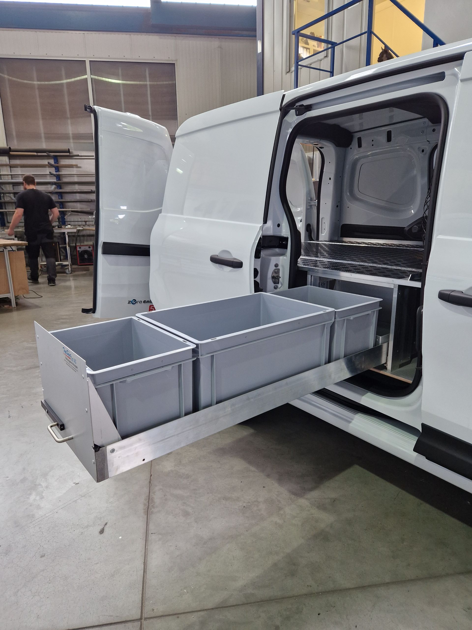 Weißer Lieferwagen mit geöffneter Seitentür, hinter der sich ausziehbare Lagerbehälter befinden. Graue Behälter auf einem Metallrahmen in einer Industrieumgebung.