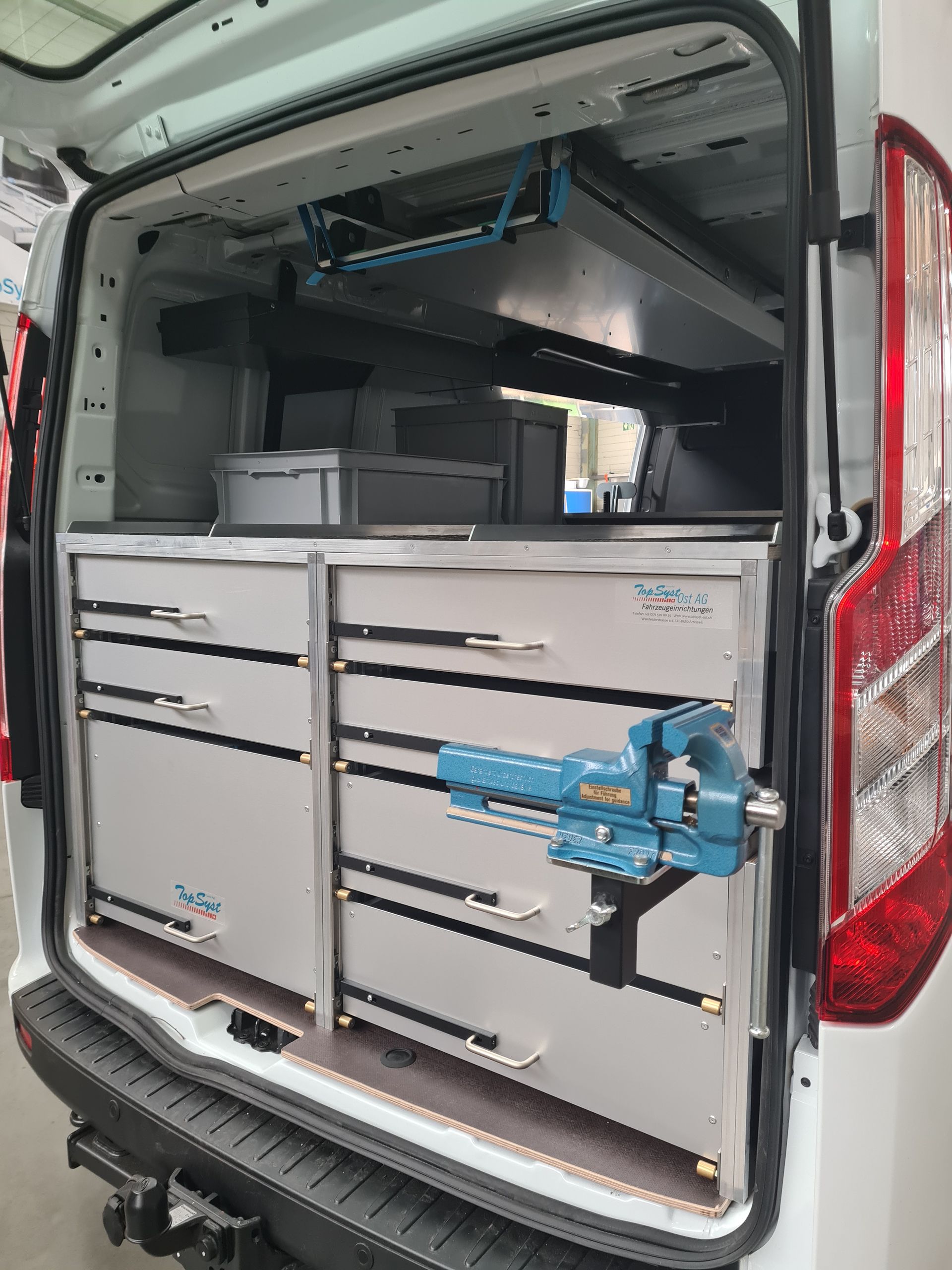 Innenansicht eines weißen Arbeitswagens mit Metallschubladen, Stauraum und einem seitlich montierten blauen Schraubstock.