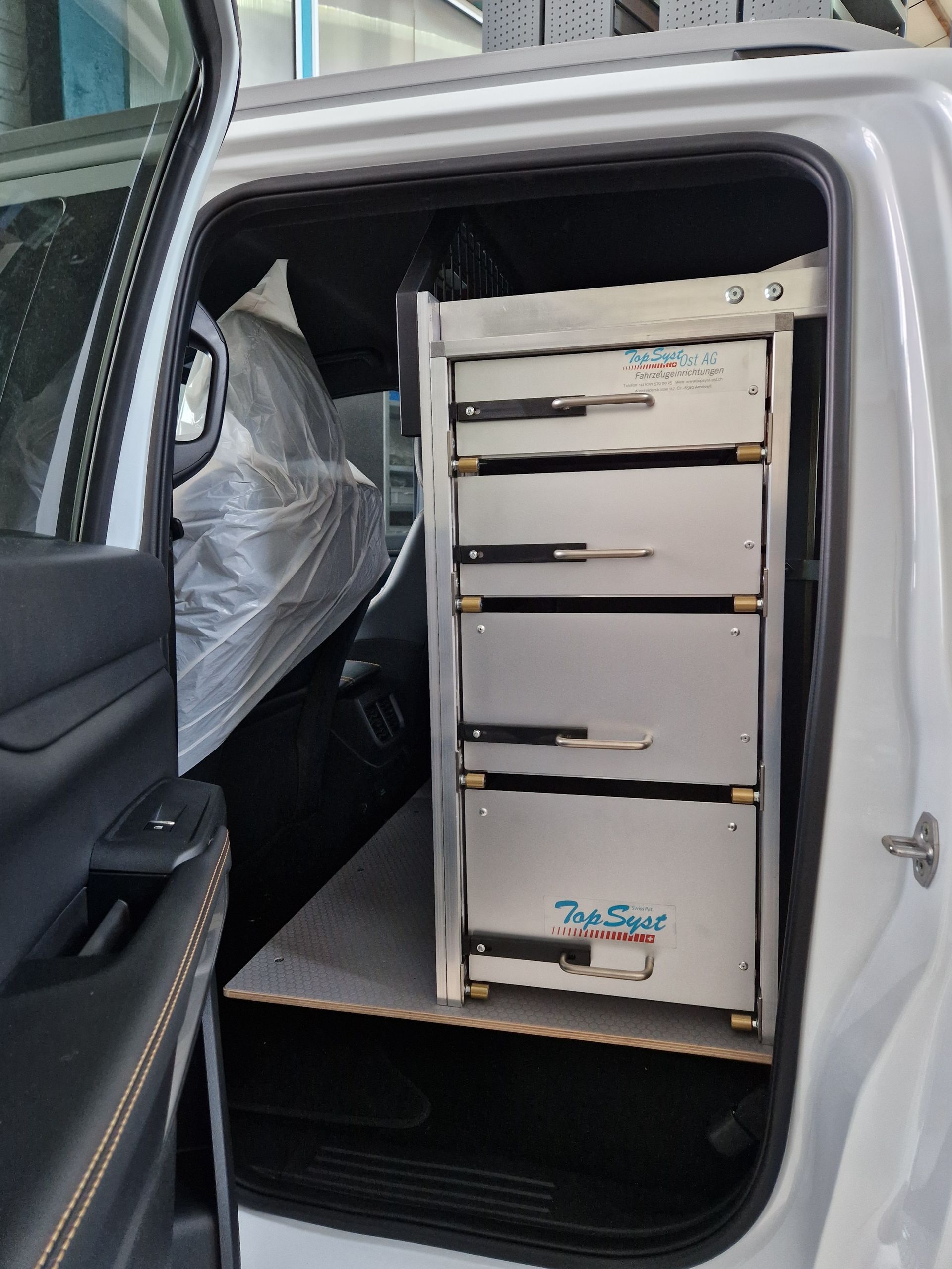 In der offenen Tür eines weißen Fahrzeugs steht ein hohes, silbernes Metallregal mit vier Schubladen.