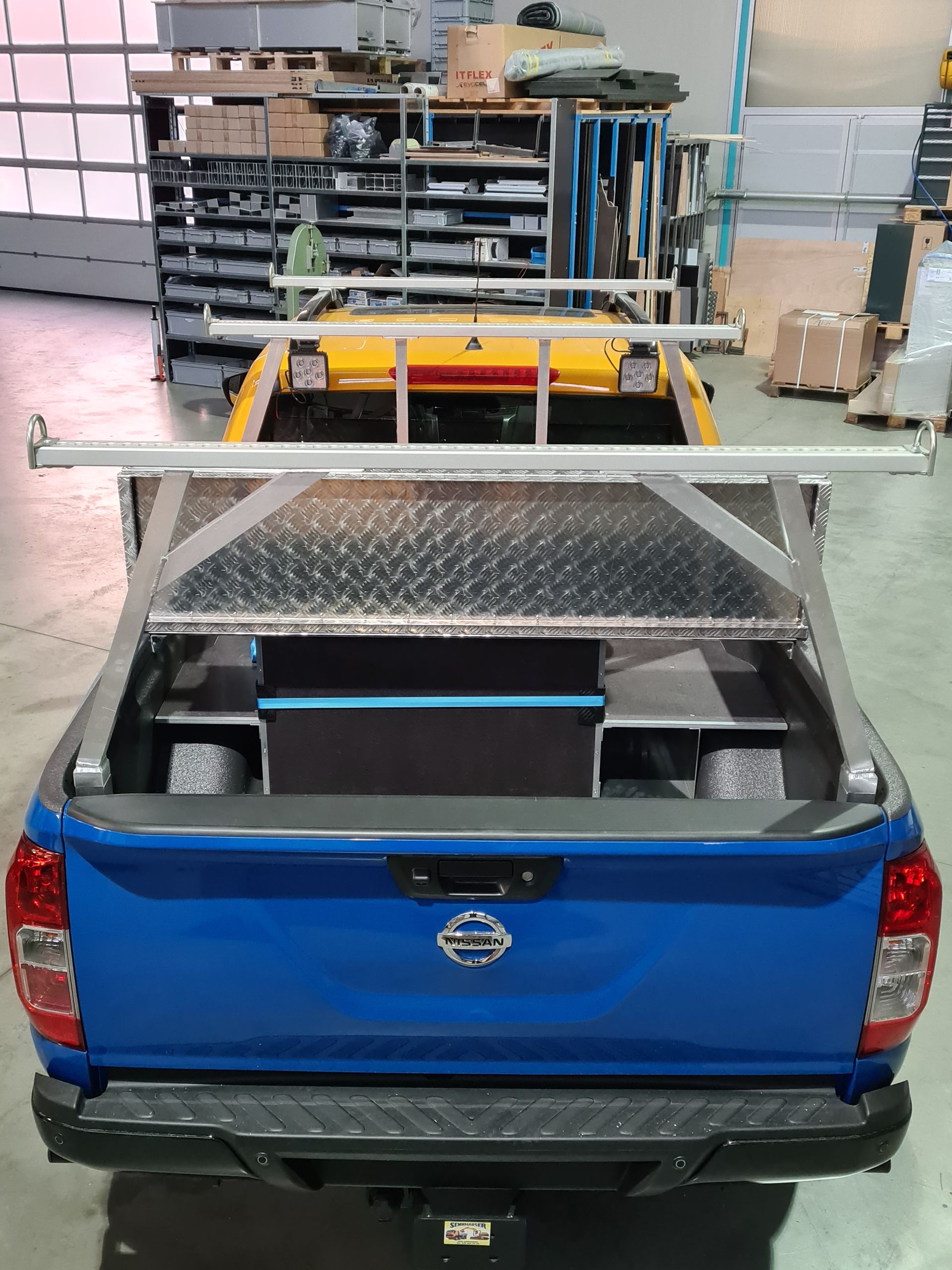 Blauer Nissan-Pickup mit Metalllagerung und -gestell, gelber Truck im Hintergrund.