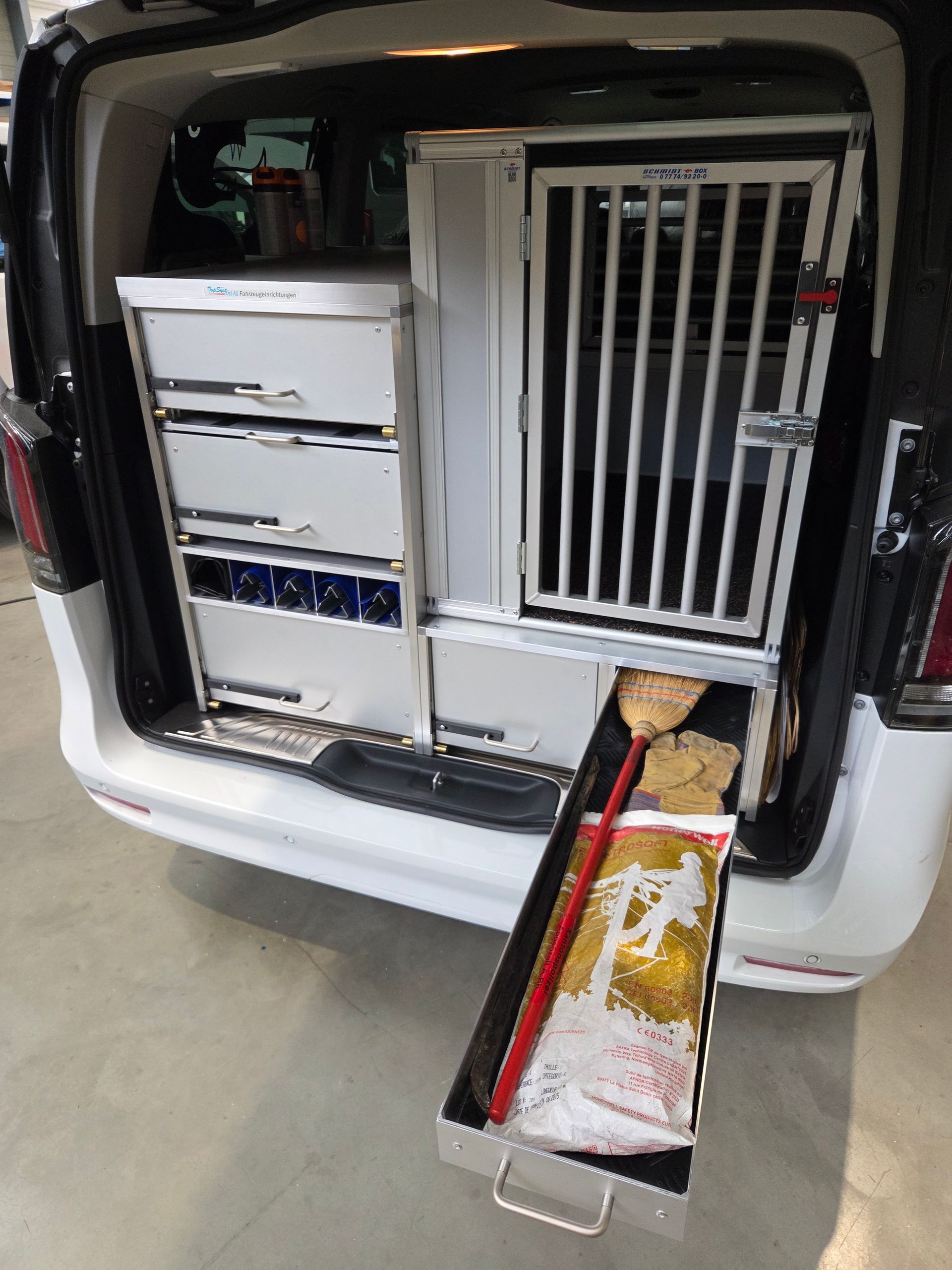 Innenraum eines weißen Lieferwagens mit einer Hundebox und Lagerschränken; in einer Schublade befinden sich ein Besen und eine Fast-Food-Tüte.
