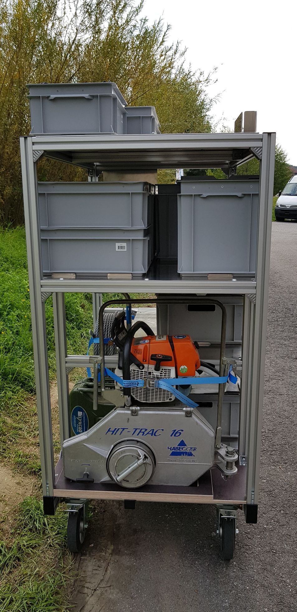 Ein Metallwagen mit Rädern, grauen Lagerbehältern und einer Kettensäge. Im Freien mit Bäumen und einer Straße im Hintergrund.