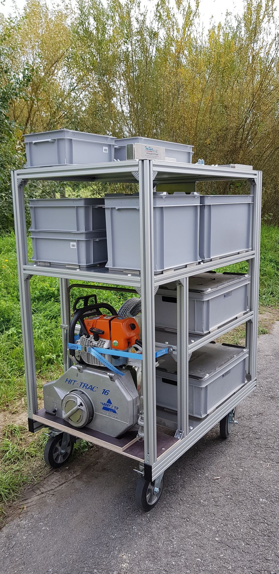 Ein Wagen mit Regalen und grauen Behältern. Auf dem unteren Regal im Freien stehen ein Motor und eine Kettensäge.