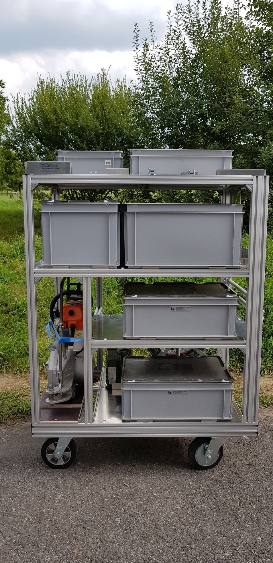 Ein Metallwagen mit grauen Lagerbehältern, Rädern und Maschinen auf einer Rasenfläche mit Bäumen.