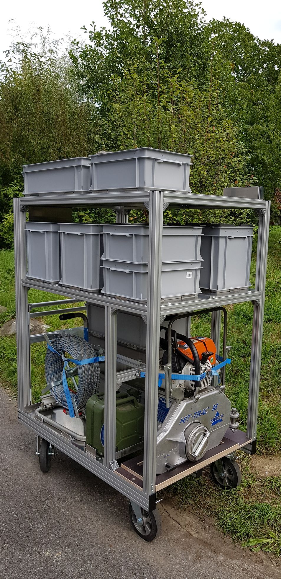 Ein Metallkarren mit Kisten und Maschinen auf einer gepflasterten Fläche vor einem Hintergrund aus Bäumen.