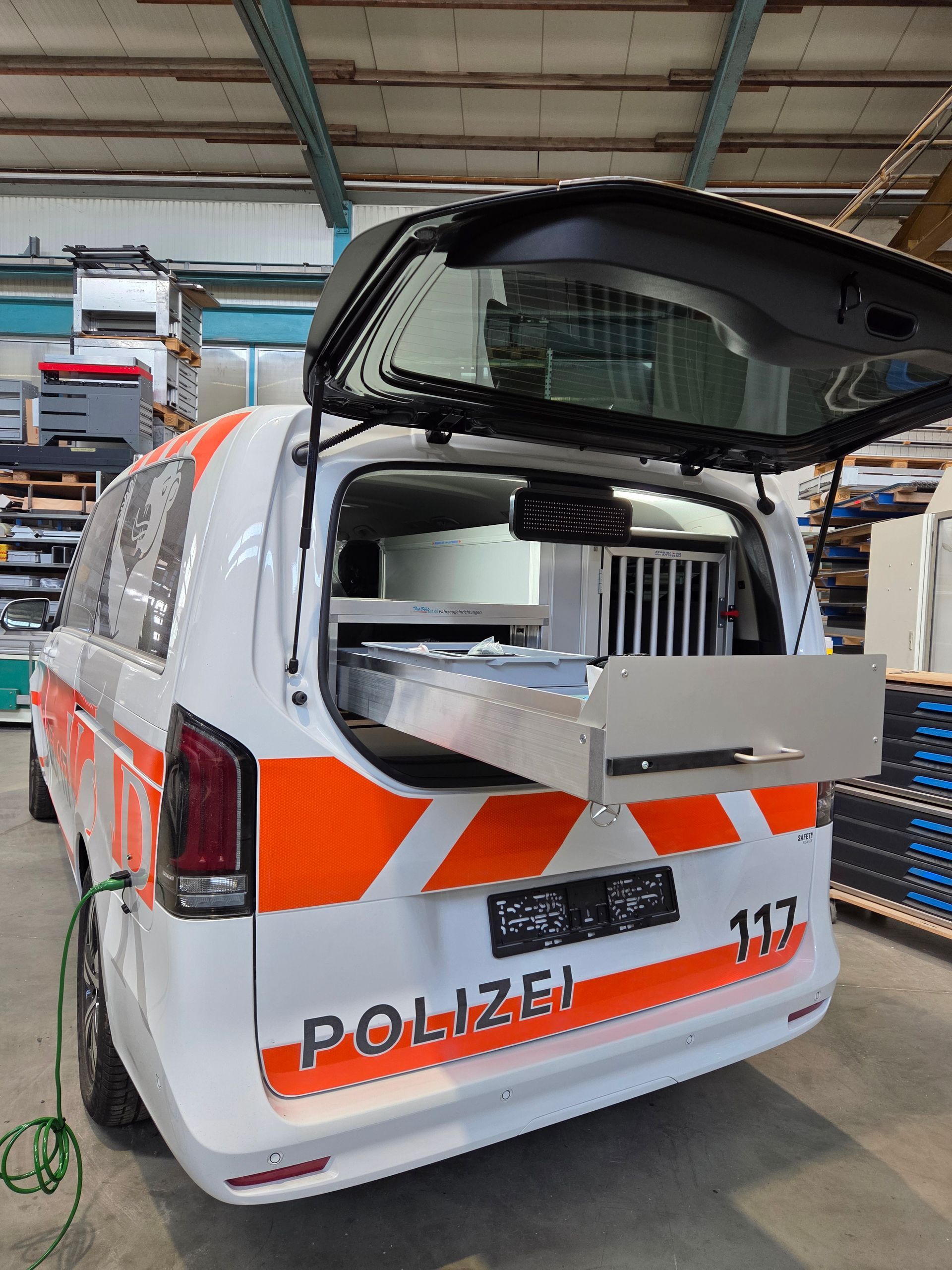 Rückansicht eines weiß-orangen Polizeiwagens mit geöffnetem Kofferraum, der eine ausziehbare Schublade freigibt.