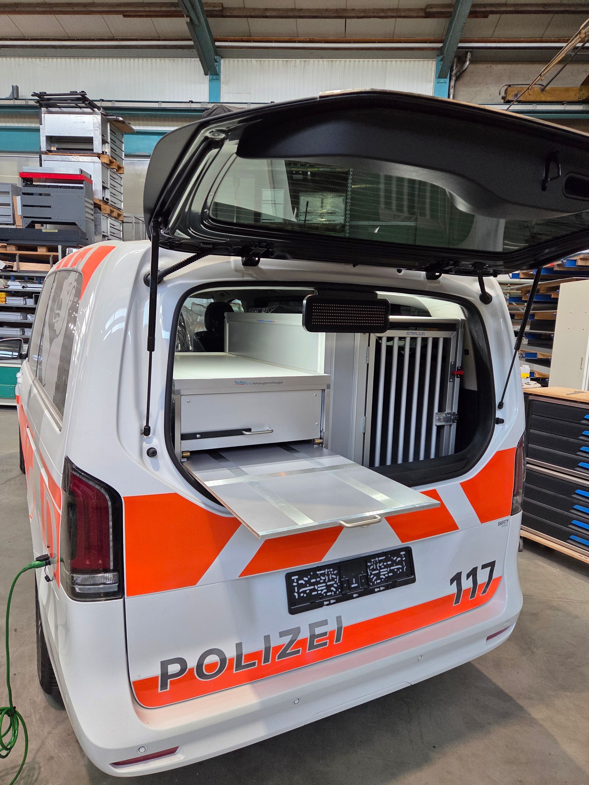 Weißer Polizeiwagen mit orangefarbenen Streifen, offene Rückseite, mit Sicht auf einen Lagerbereich. „POLIZEI“-Schriftzug sichtbar.