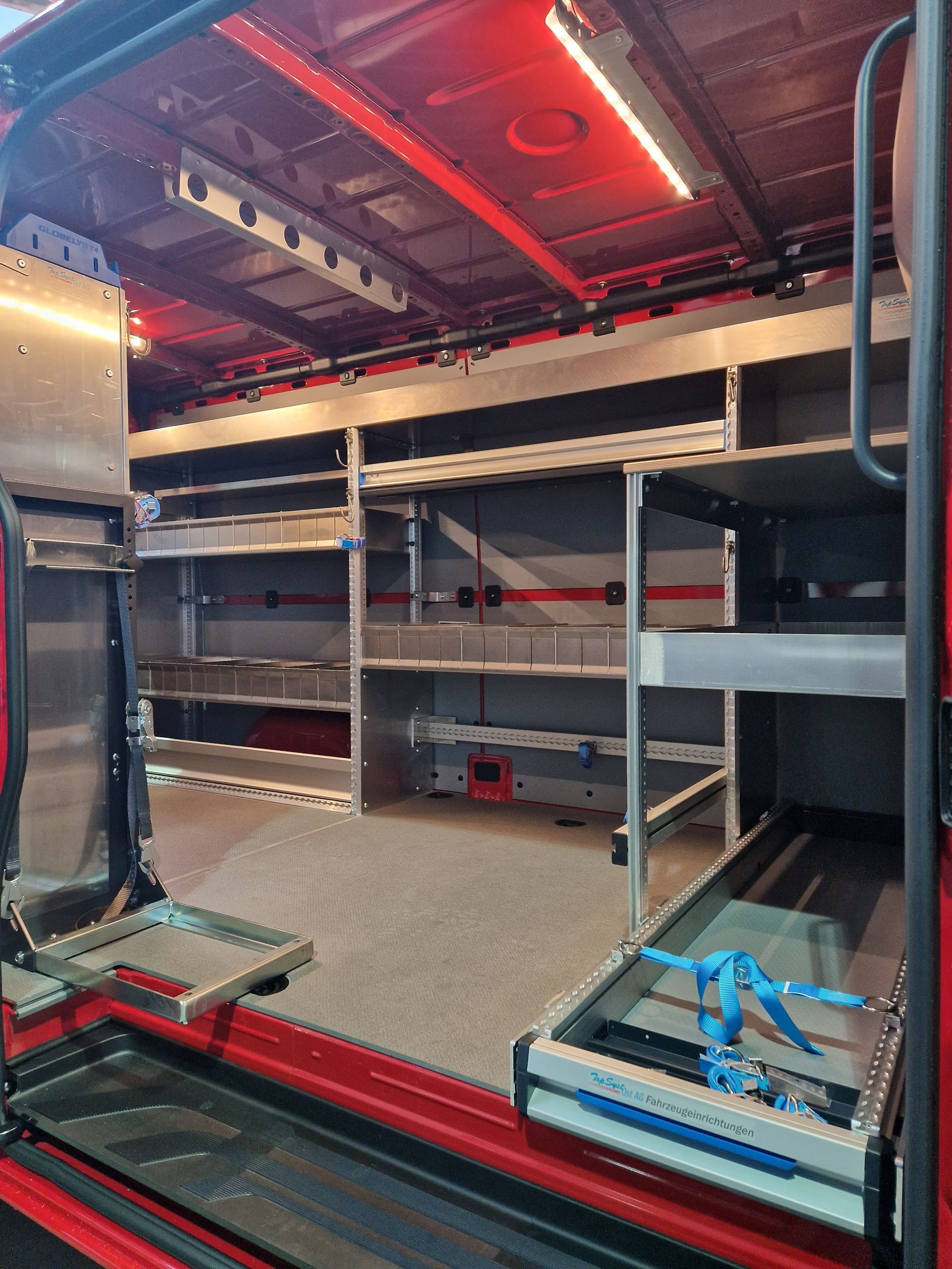Innenraum eines roten Fahrzeugs mit Regalen und Gerätelagerung. Helle Deckenbeleuchtung.