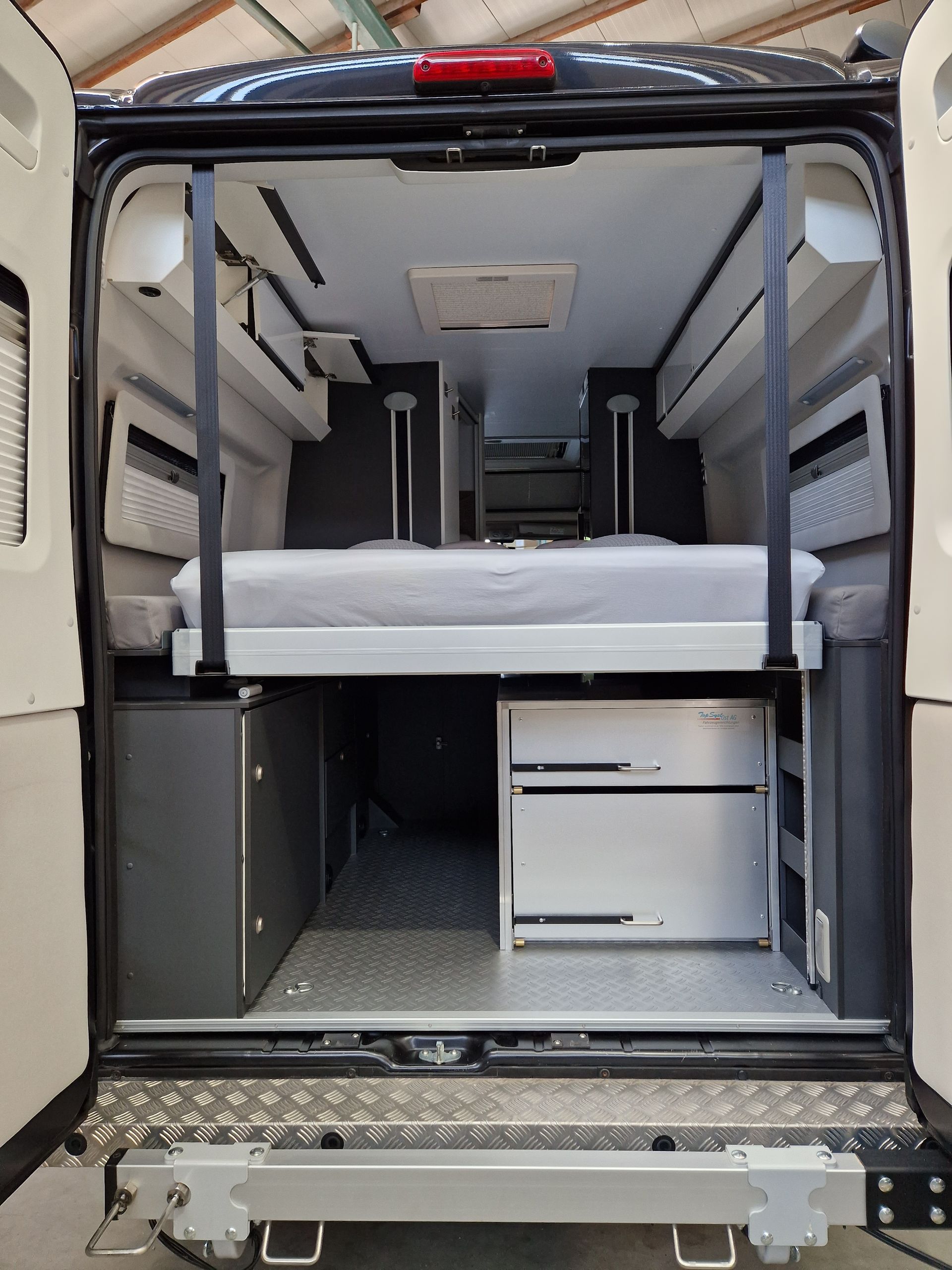 Innenansicht eines Wohnmobils mit Bett, Schränken und Metallboden, gesehen von den offenen Hintertüren.