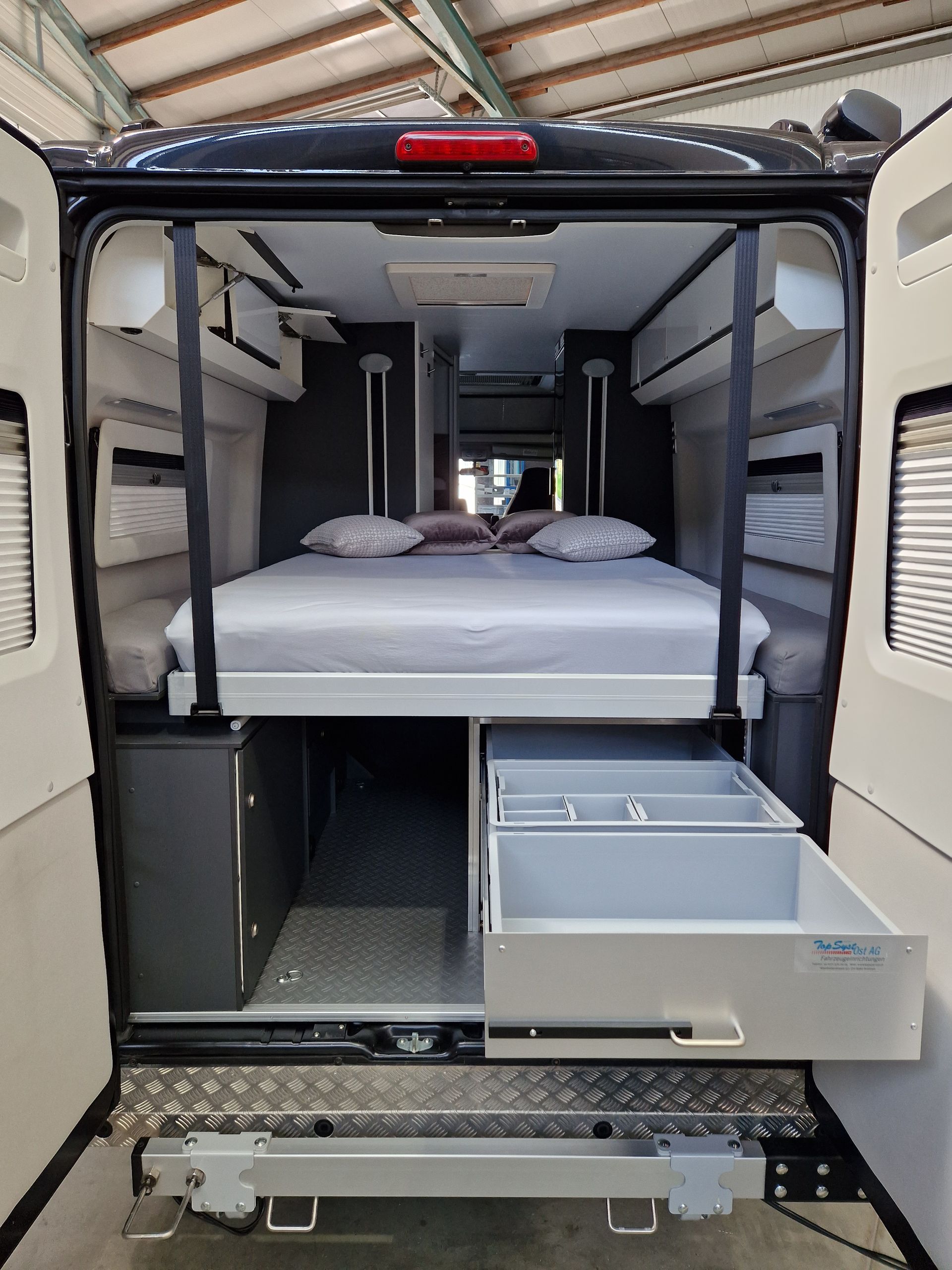 Innenansicht eines Wohnmobils mit Bett, Schränken und Schubladen. Die Farben Schwarz, Grau und Weiß dominieren.