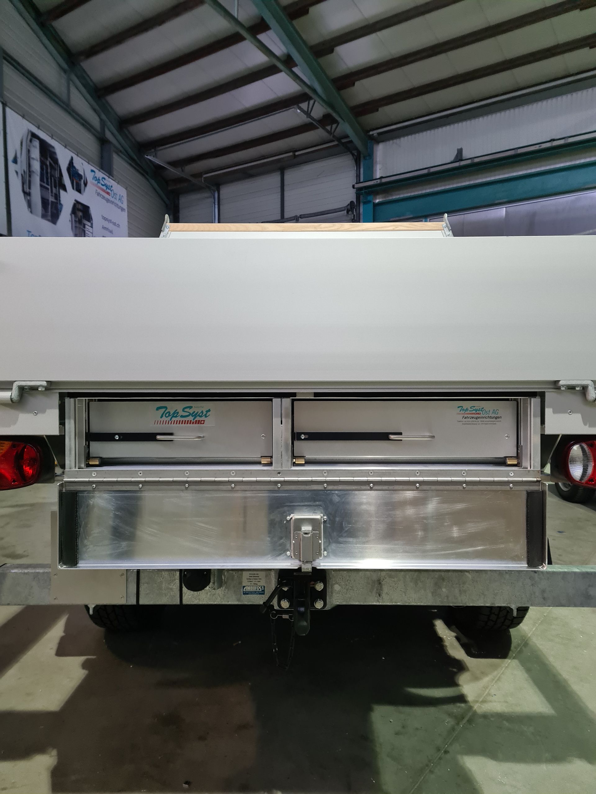 Rückansicht eines silbernen Anhängers mit zwei Schubladen, einer Anhängerkupplung und roten Rücklichtern in einer Lagerhalle.