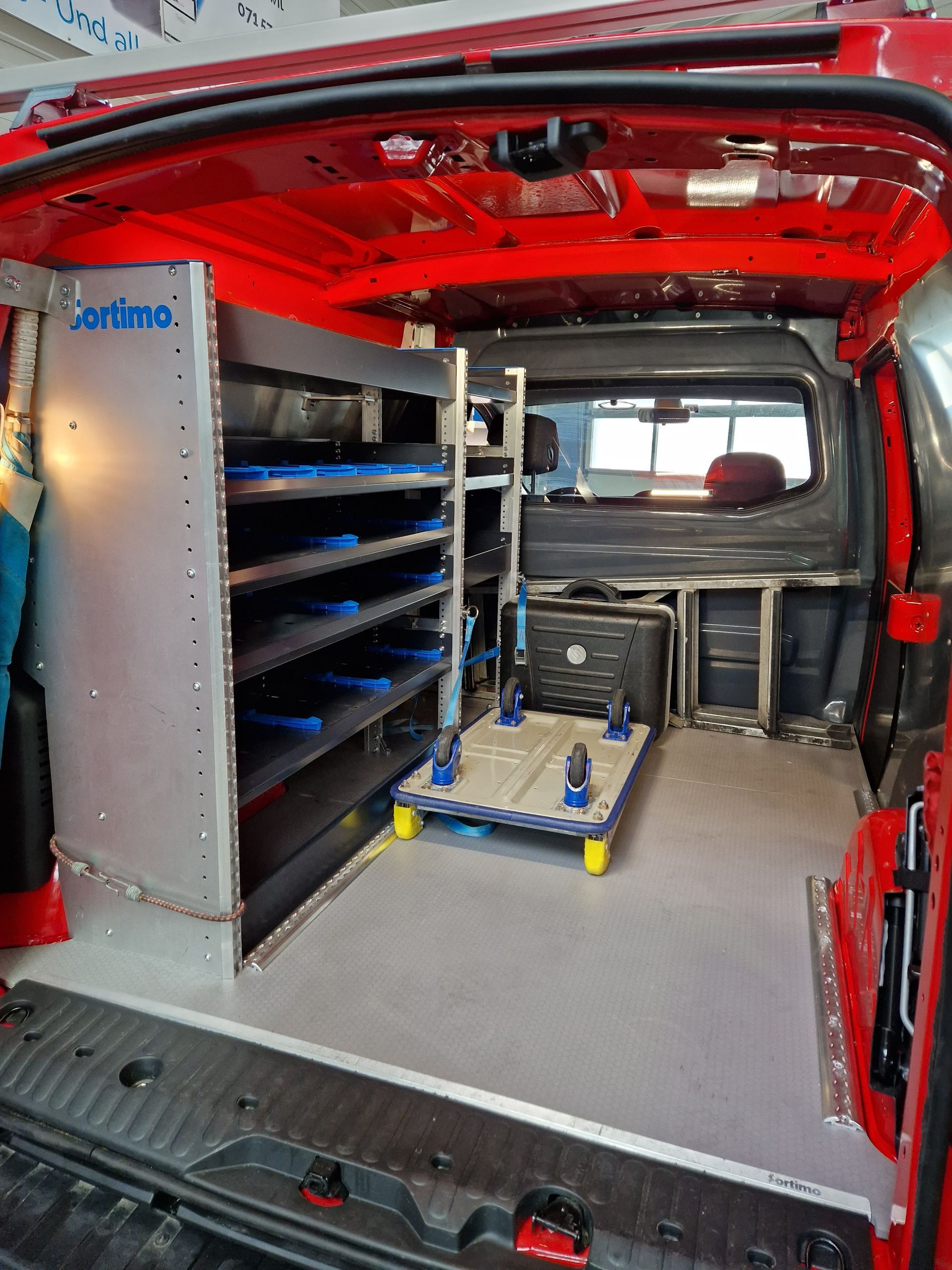 Innenraum eines roten Lieferwagens mit Regalen, Werkzeugwagen und grauem Boden.