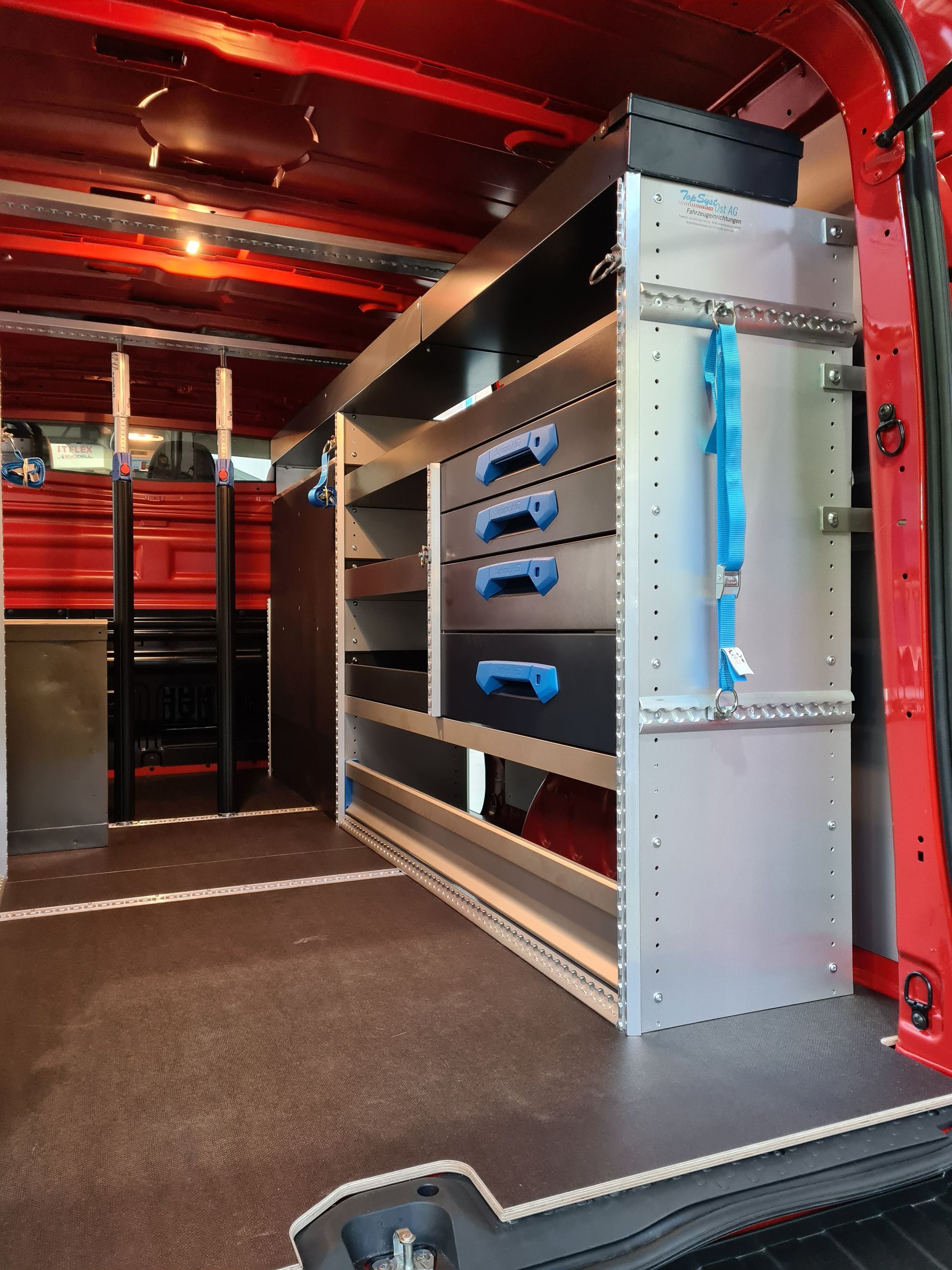 Innenraum eines roten Arbeitstransporters, ausgestattet mit Metallregalen und Schubladen für Werkzeuge, auf einem schwarzen Boden.