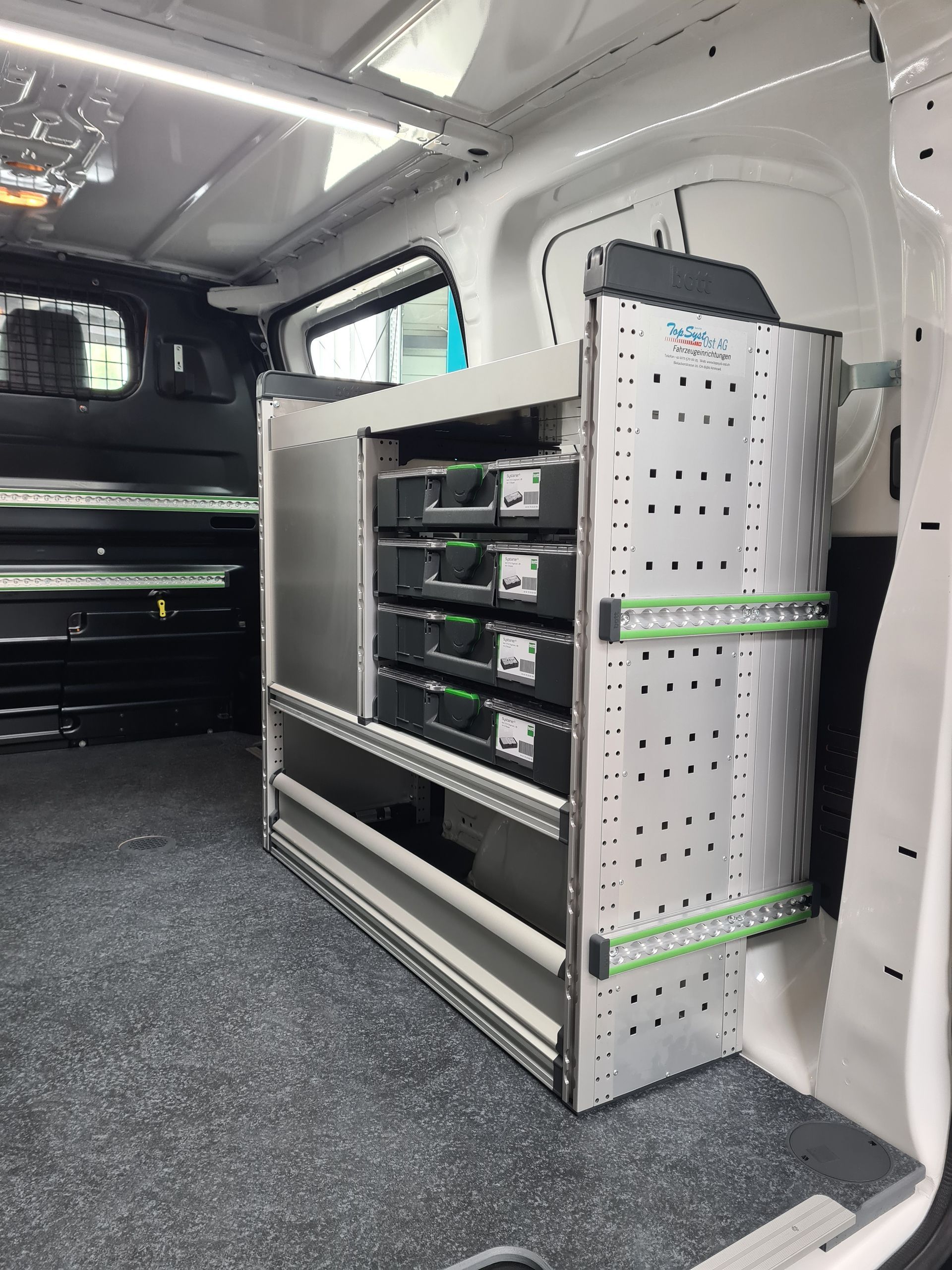 Innenraum eines Lieferwagens mit Regalen und Stauraum. Die Regale sind aus Metall mit grünen Akzenten. Der Boden ist dunkelgrau.