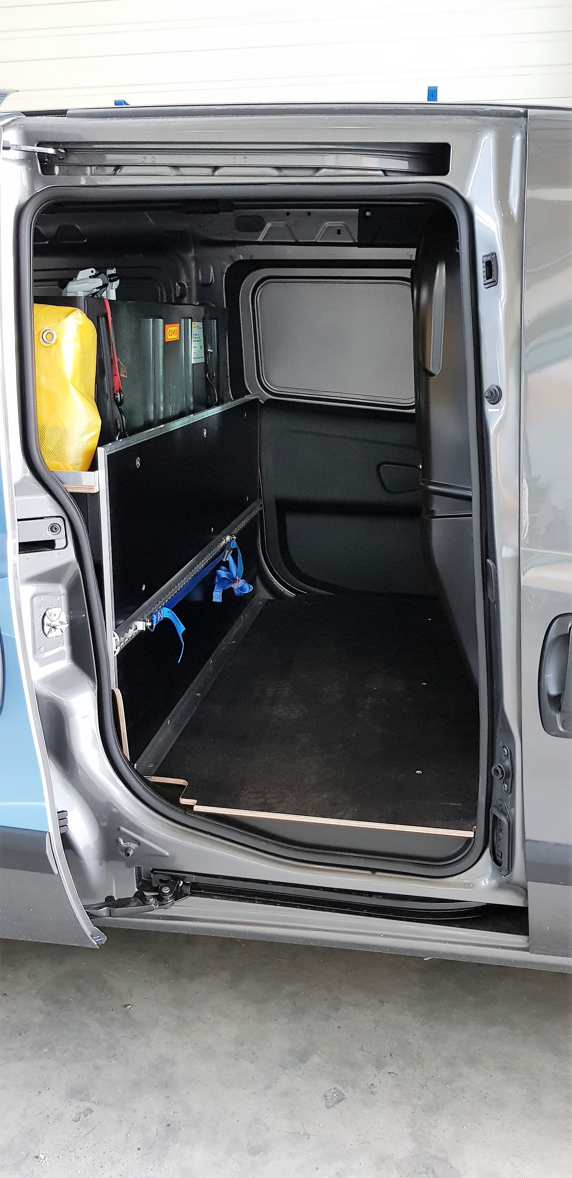 Hinter der offenen Seitentür eines grauen Lieferwagens sind maßgefertigte Regale und Lagerräume zu sehen; ein gelber Container ist sichtbar.