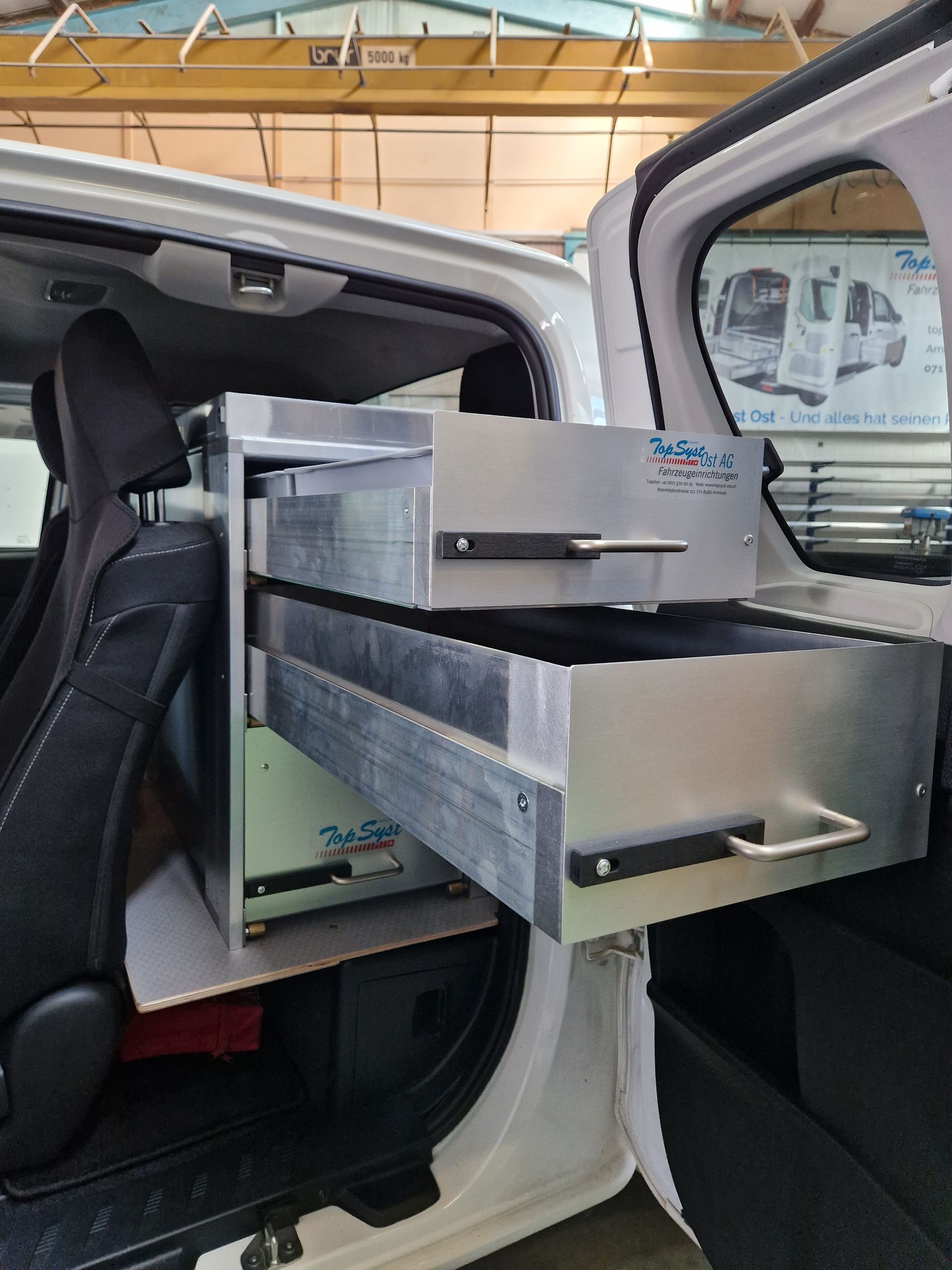 Weißer Lieferwageninnenraum mit offener Metallschublade neben einem schwarzen Sitz; Werkstatt-Setting.