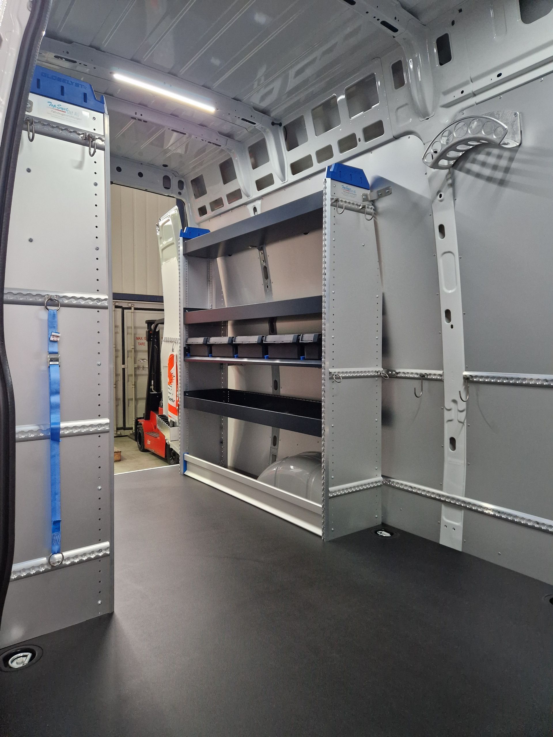 Innenraum eines weißen Lieferwagens mit grauen Regalen, schwarzem Boden und einem im Hintergrund sichtbaren Gabelstapler.