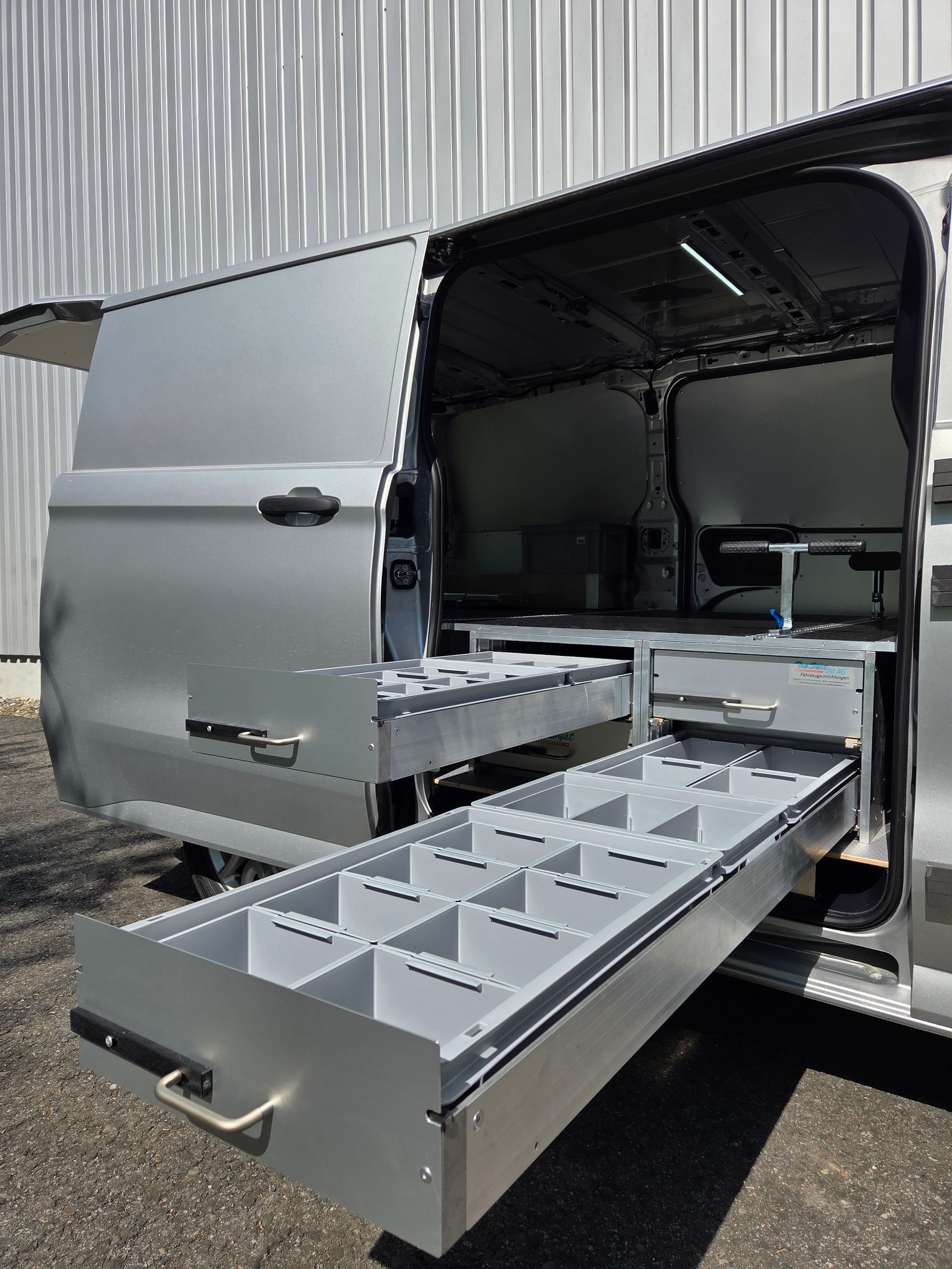 Silberner Lieferwagen mit offener Hecktür, hinter der sich ausziehbare Schubladen mit Unterteilungen für Werkzeuge befinden.