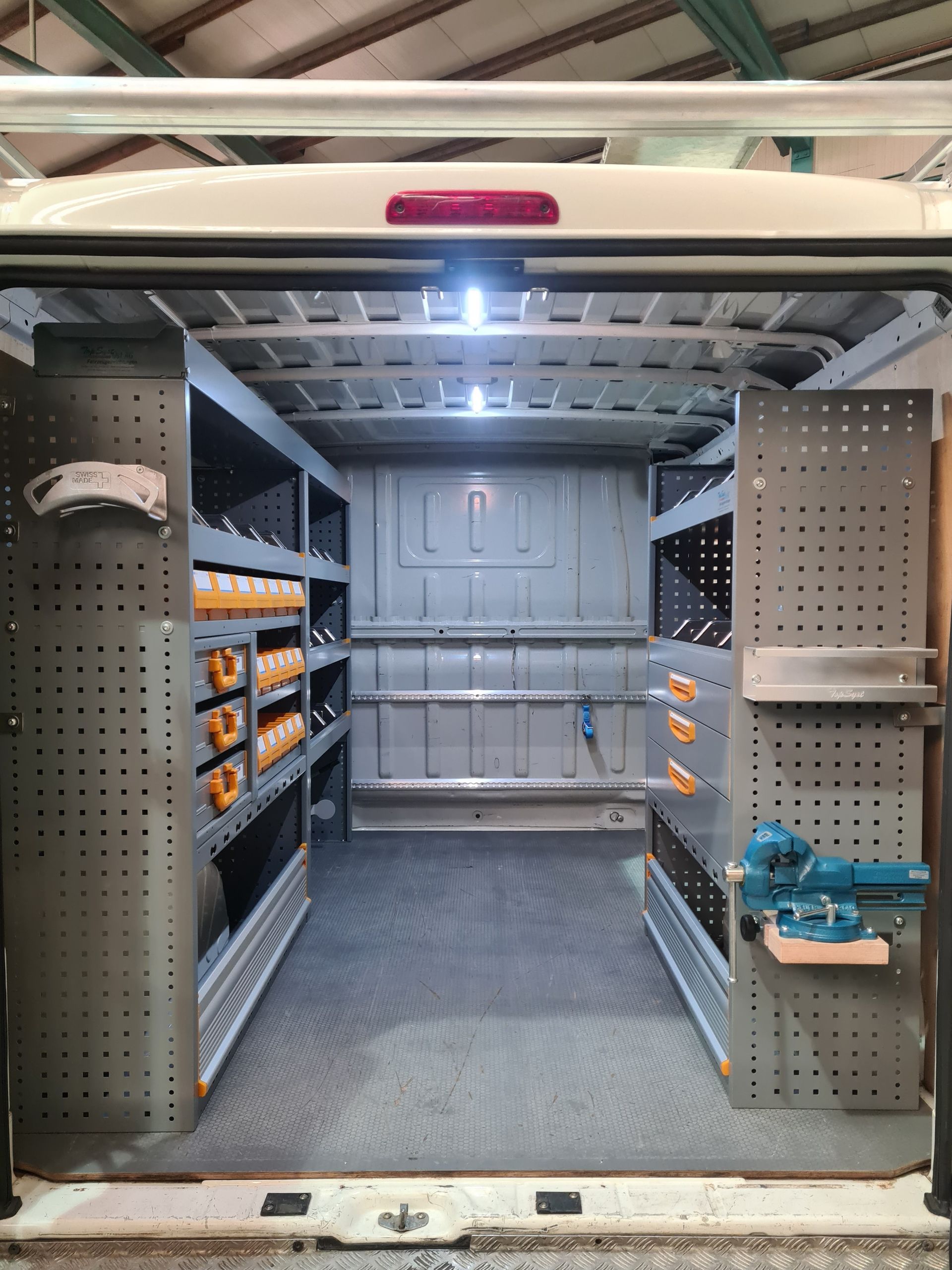 Innenraum eines weißen Arbeitstransporters mit Metallregalen, Lagerbehältern und Werkzeugen, für den Transport organisiert.