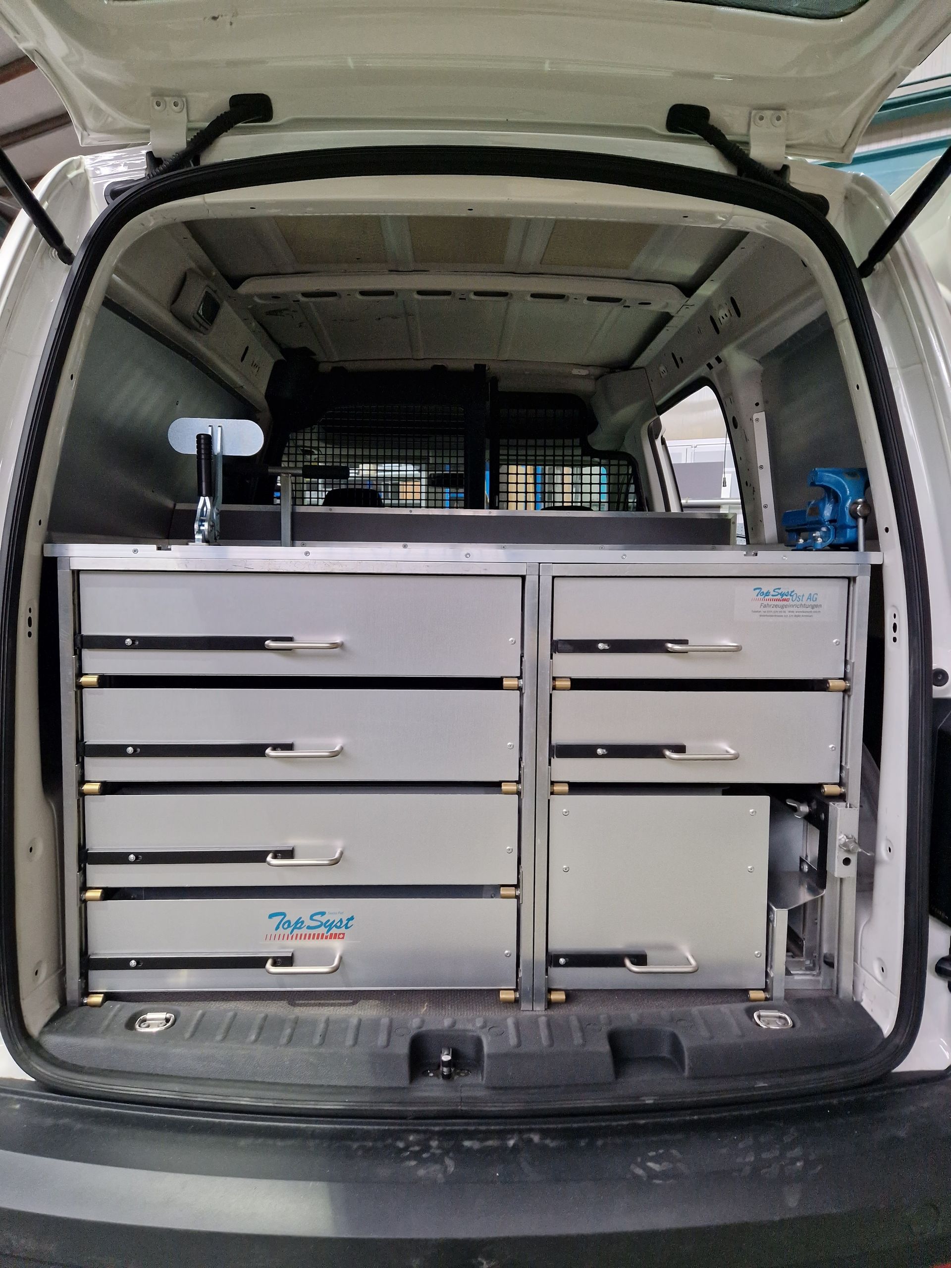 Innenansicht eines weißen Lieferwagens mit maßgefertigten Metallschubladen und einem Arbeitsbereich, wahrscheinlich für Werkzeuge, an der Hintertür.
