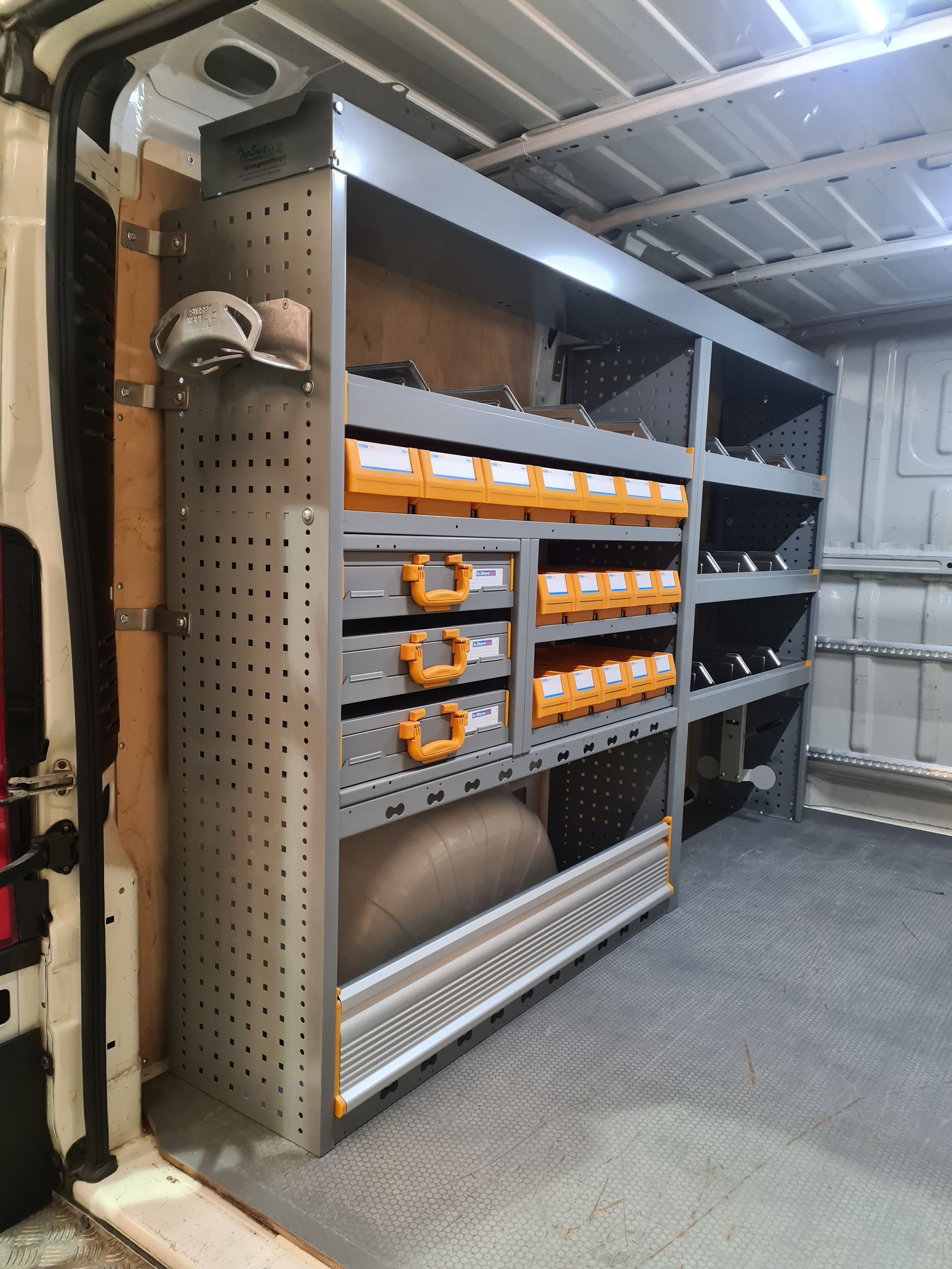 Innenausstattung des Lieferwagens mit Metallregal, orangefarbenen Behältern, Schubladen, grauen und silbernen Werkzeugen.