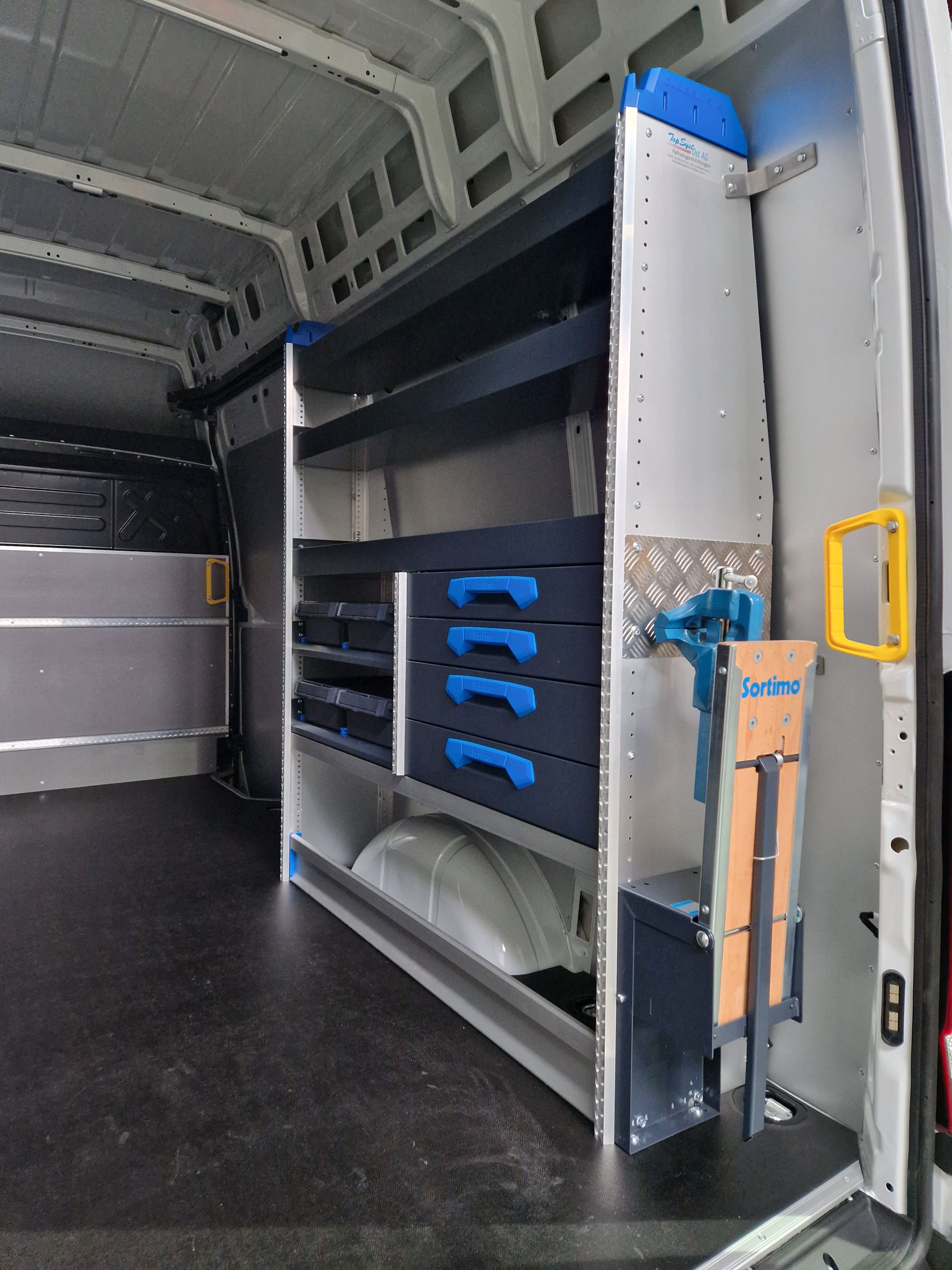 Innenraum eines weißen Lieferwagens mit Regaleinheit; blaue Schubladen, Schraubstock und seitlich montierte Werkzeuge.