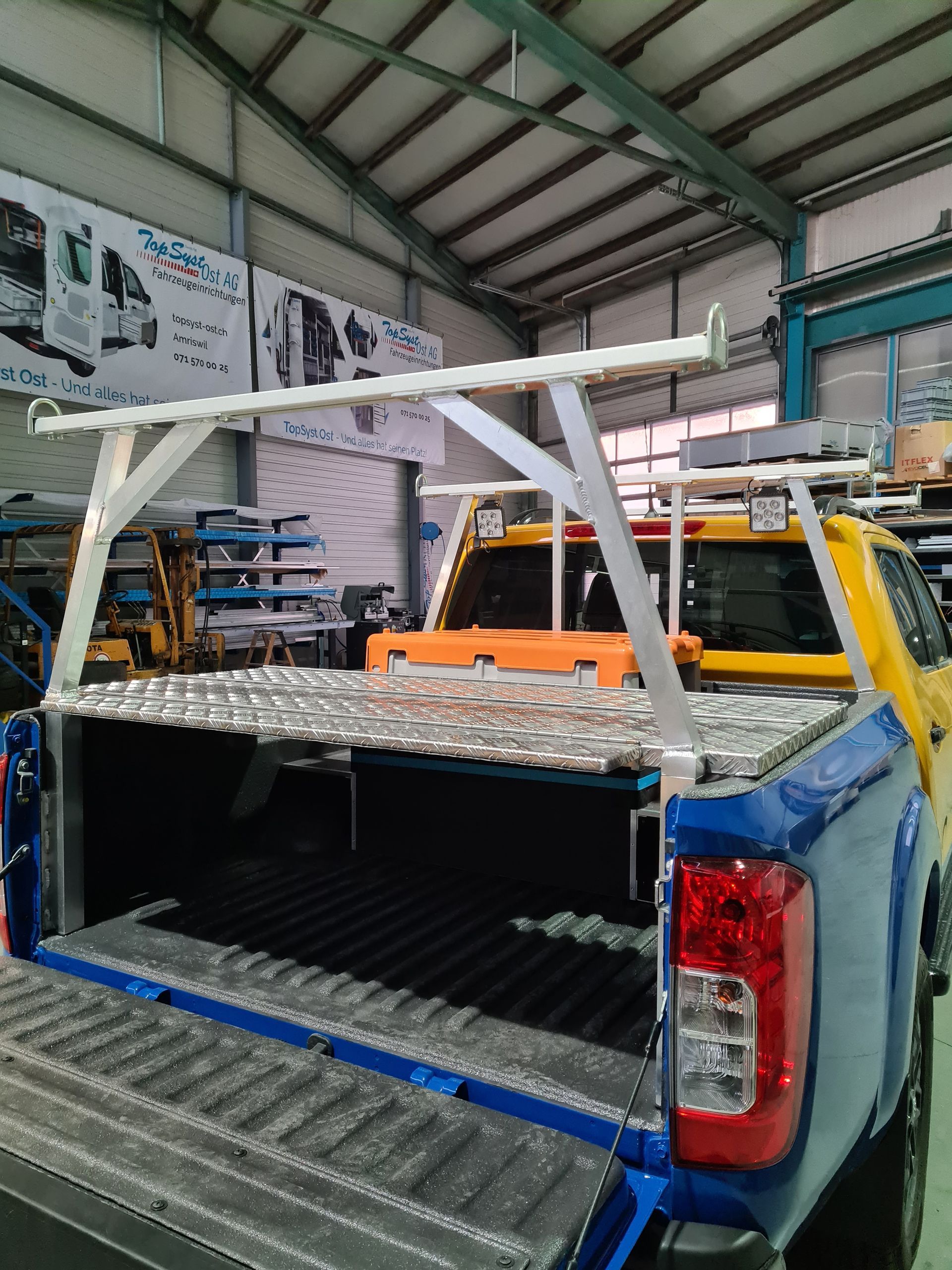 Ein blauer Pickup mit silberner Ladeflächenabdeckung und Leitergestell in einem Lagerhaus.