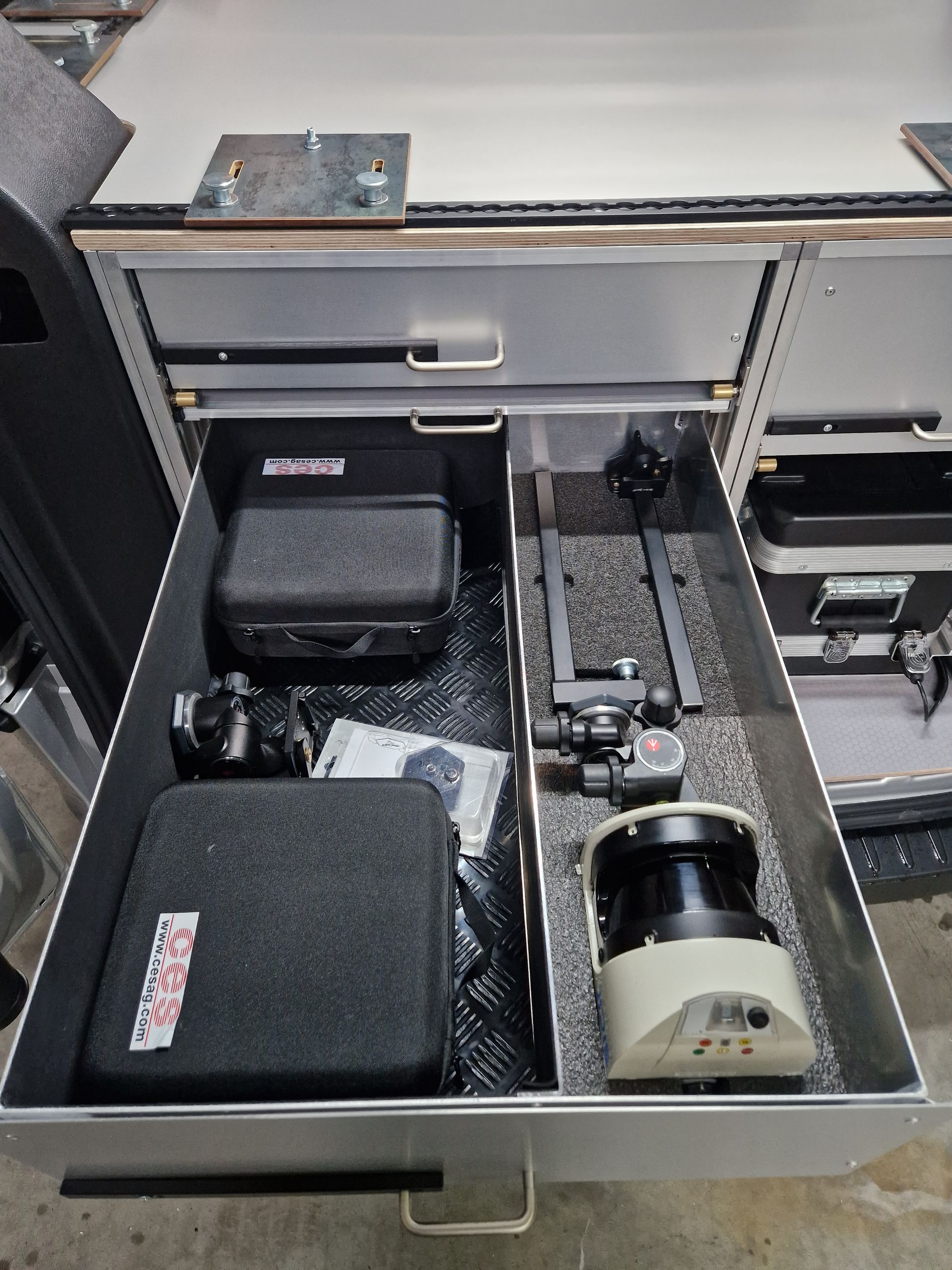 Offene Schublade mit Vermessungsgeräten, darunter Koffer, ein Stativ und ein Laserscanner.