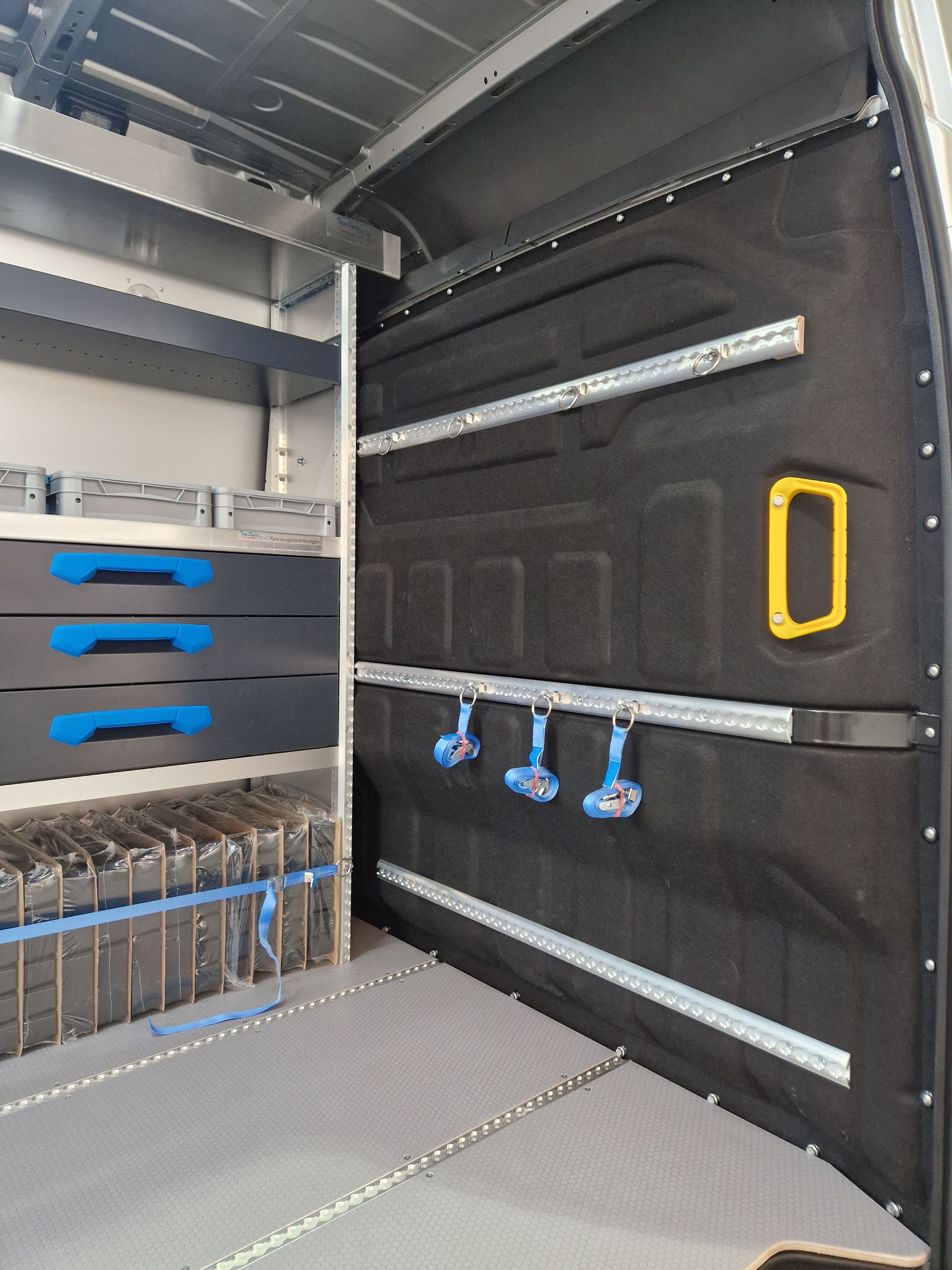 Innenraum eines Arbeitswagens mit Regalen, Schubladen und Geräteaufbewahrung. Schwarzes Wandpaneel mit Aufhängehaken.