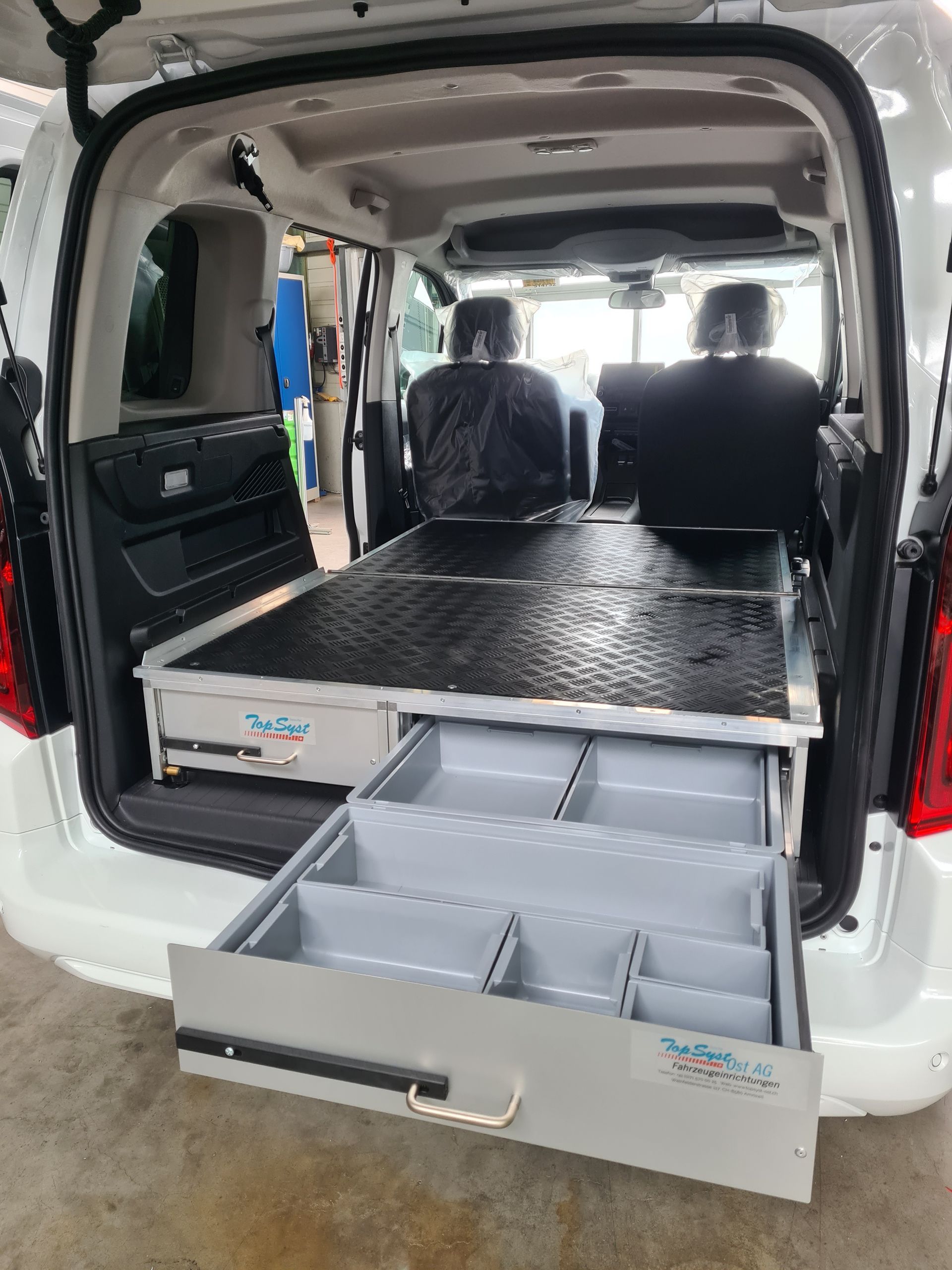 Weißer Van-Innenraum mit offenen Schubladen, grauen Trennwänden, schwarzer Plattform, sichtbaren Sitzen.