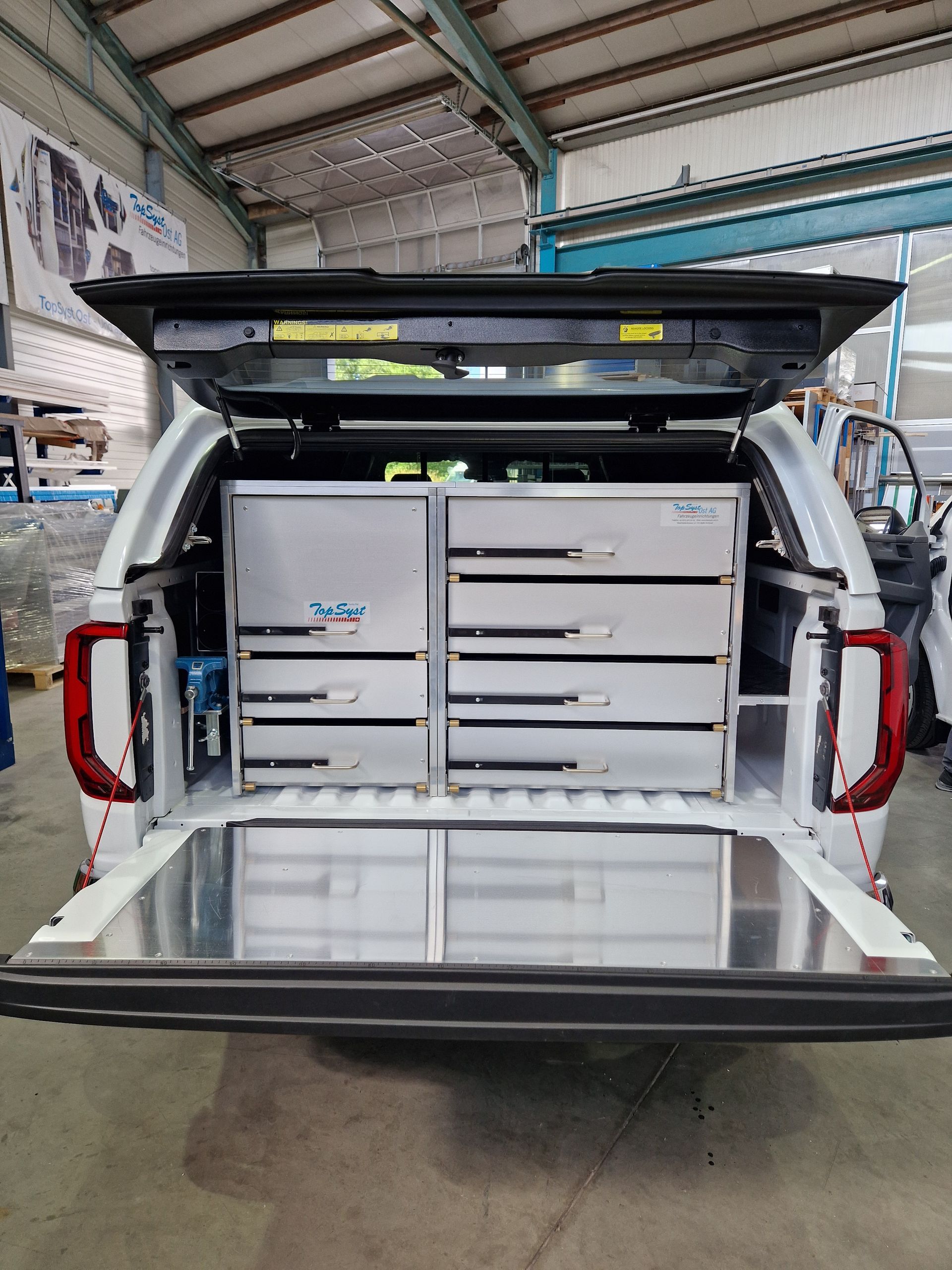 Weiße Ladefläche eines Pickups mit Schubladen, Heckklappe geöffnet. Rücklichter sind sichtbar.