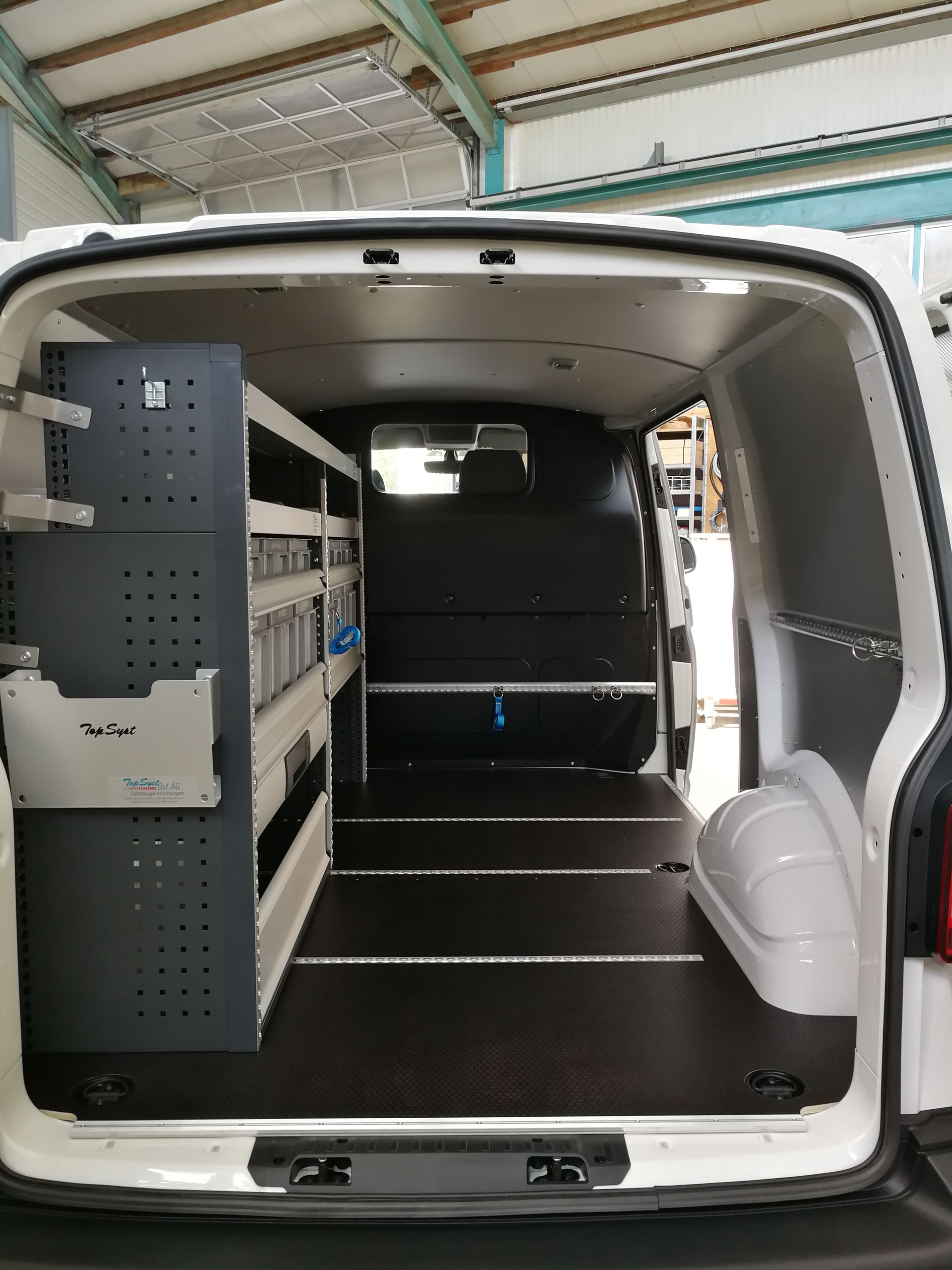 Innenraum eines weißen Lieferwagens mit Regalen, dunklem Boden und geöffneter Hecktür.