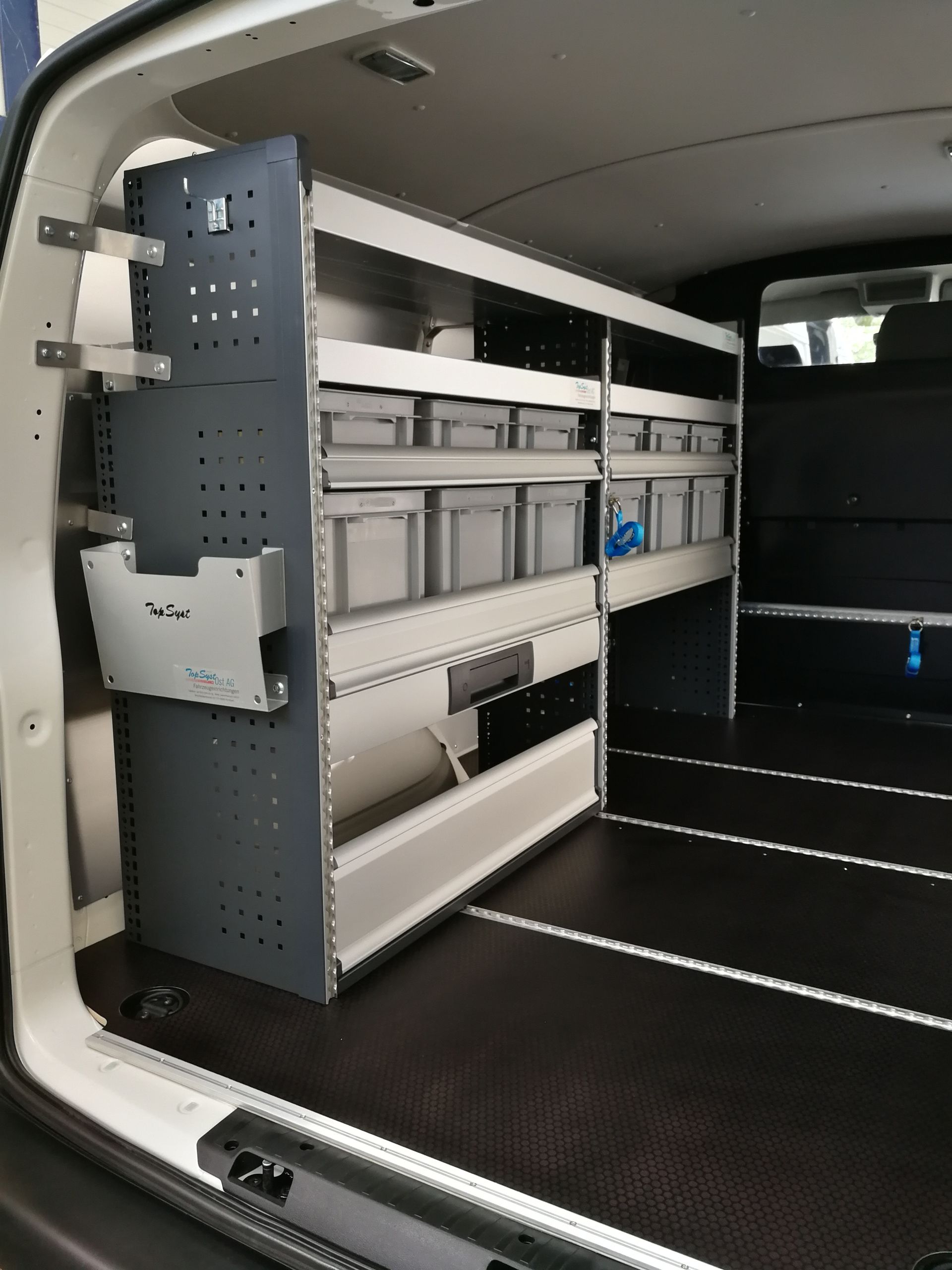 Innenraum des Lieferwagens mit Lagerregalen und -behältern; schwarzer Bodenbelag, weiße und graue Regale.