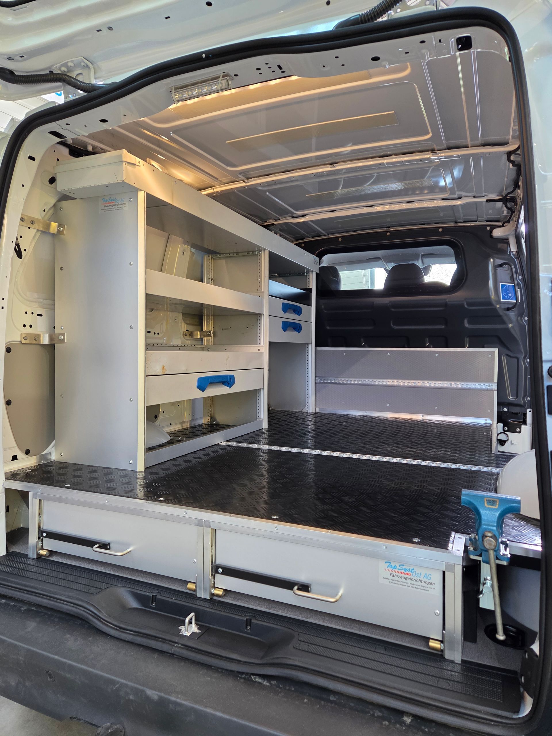 Innenraum eines weißen Lieferwagens mit Aluminiumregalen, Schubladen und einem Schraubstock zur Werkzeugaufbewahrung.