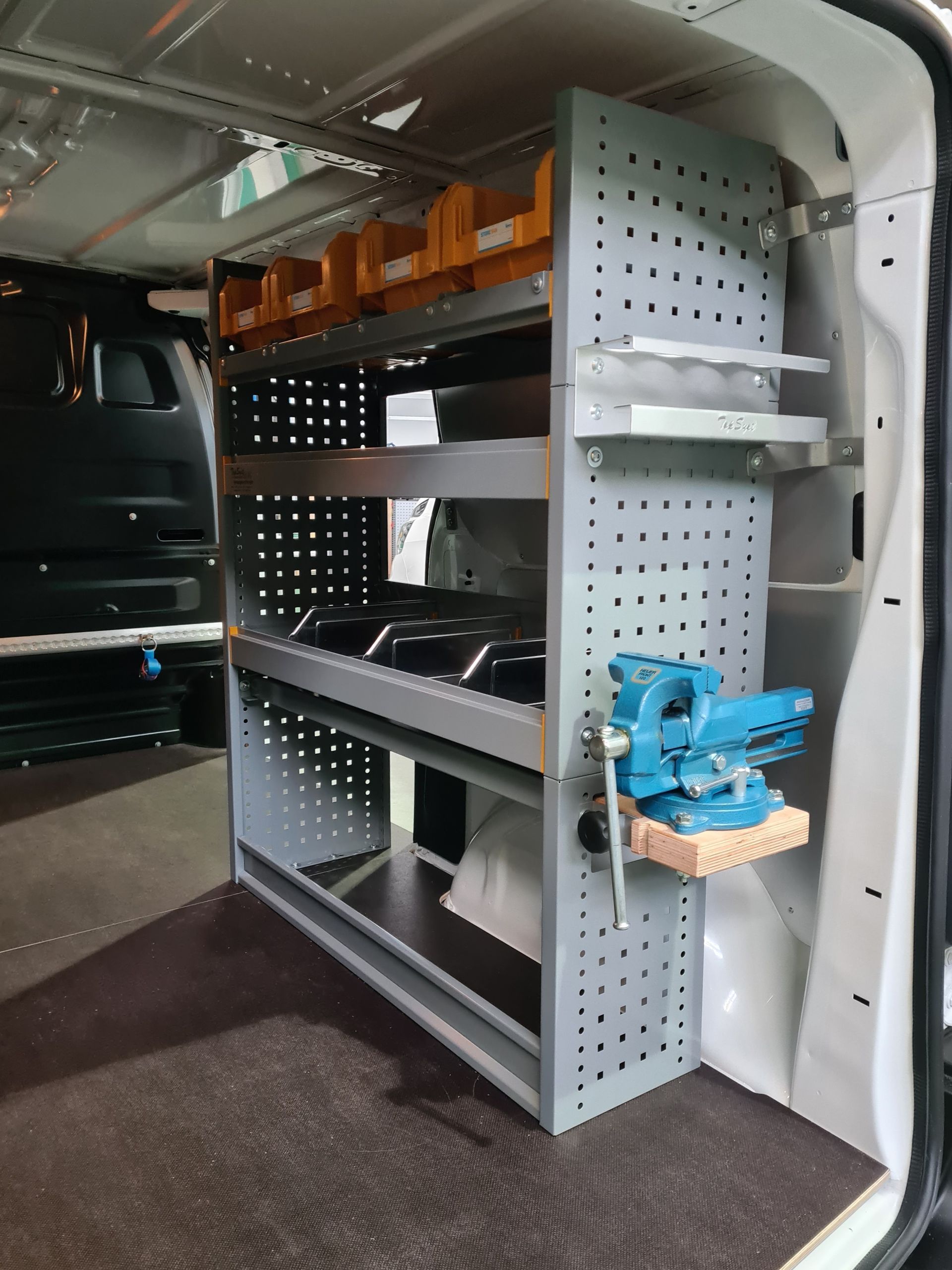 Innenraum eines Lieferwagens mit einem grauen Regal, in dem Werkzeuge und Zubehör aufbewahrt werden; ein montiertes blaues Elektrowerkzeug ist sichtbar.