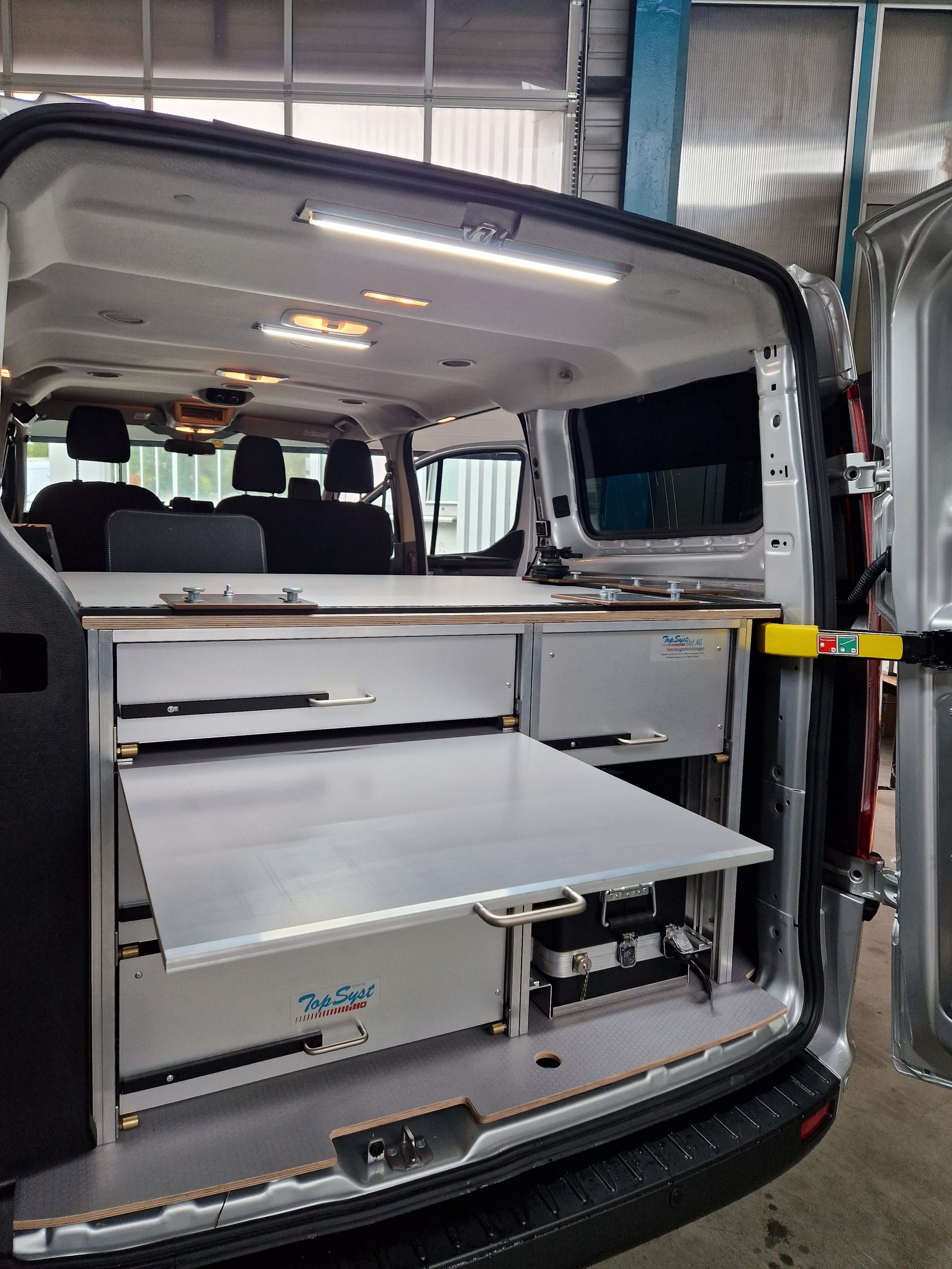 Rückansicht eines Van-Innenraums mit offenen Schubladen und einem Klapptisch. Metallische Oberflächen und LED-Beleuchtung sind sichtbar.