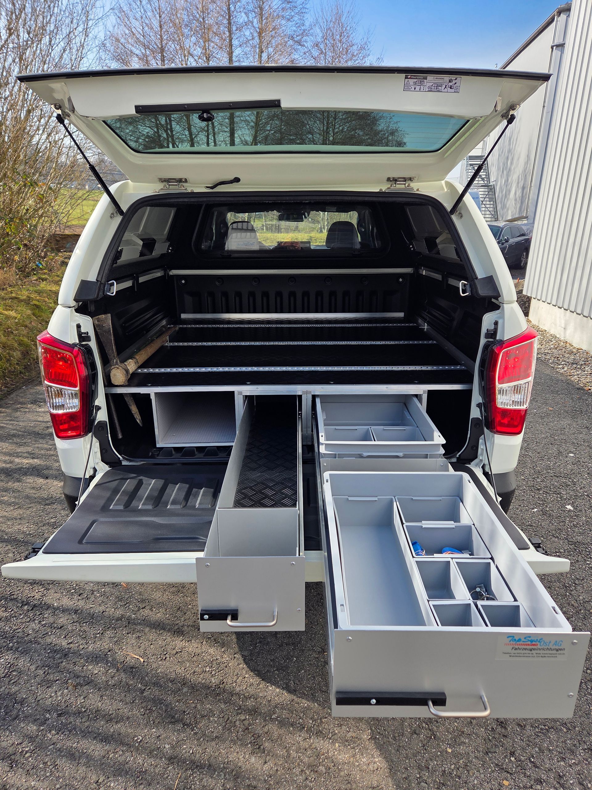 Weiße Ladefläche mit offenen Schubladen, beladen mit Werkzeug, draußen geparkt.