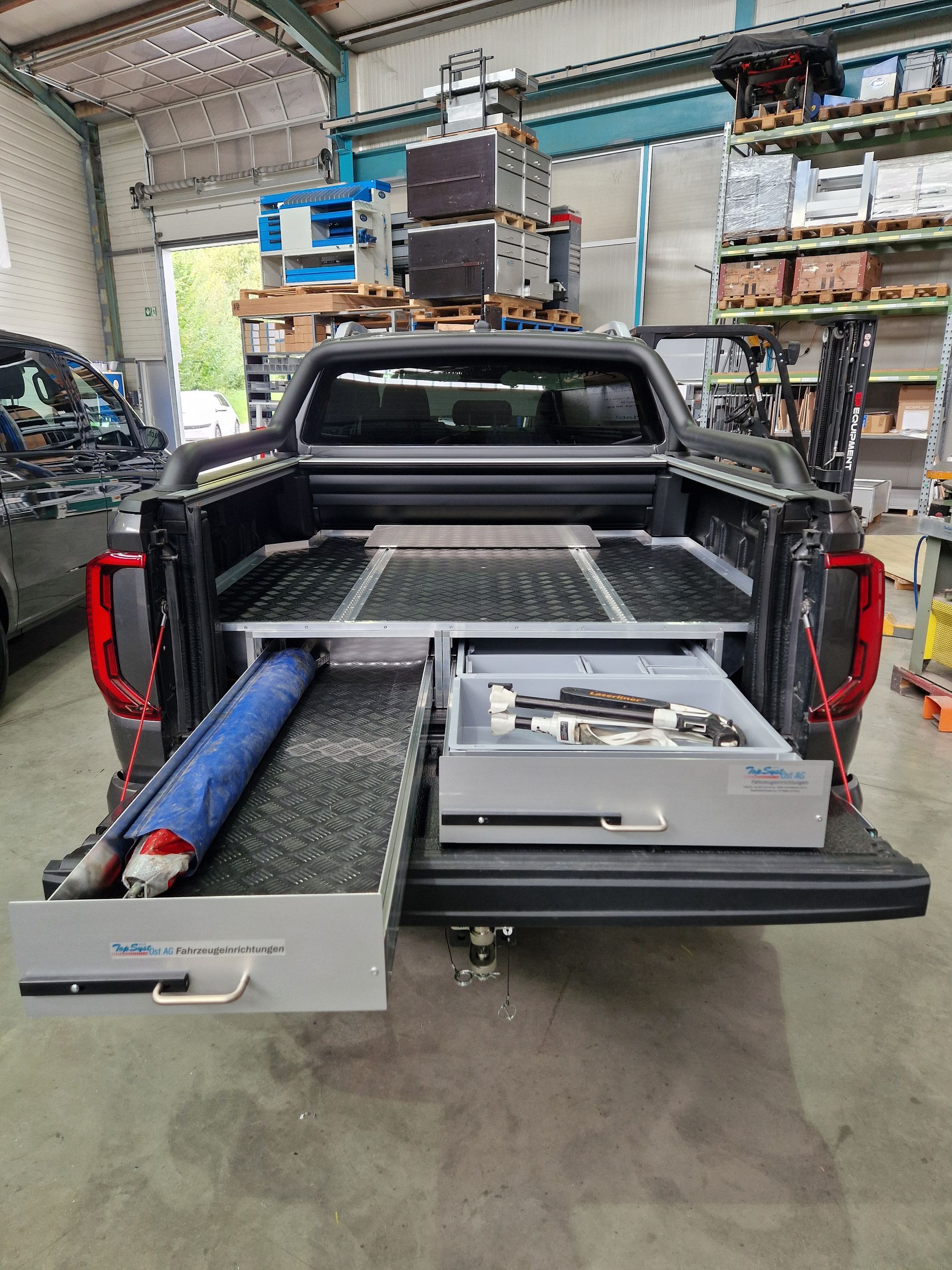 Ein schwarzer Pickup mit ausgefahrenen Schubladen. Eine enthält Werkzeuge, die andere einen zusammengerollten blauen Gegenstand. Werkstatt-Setting.