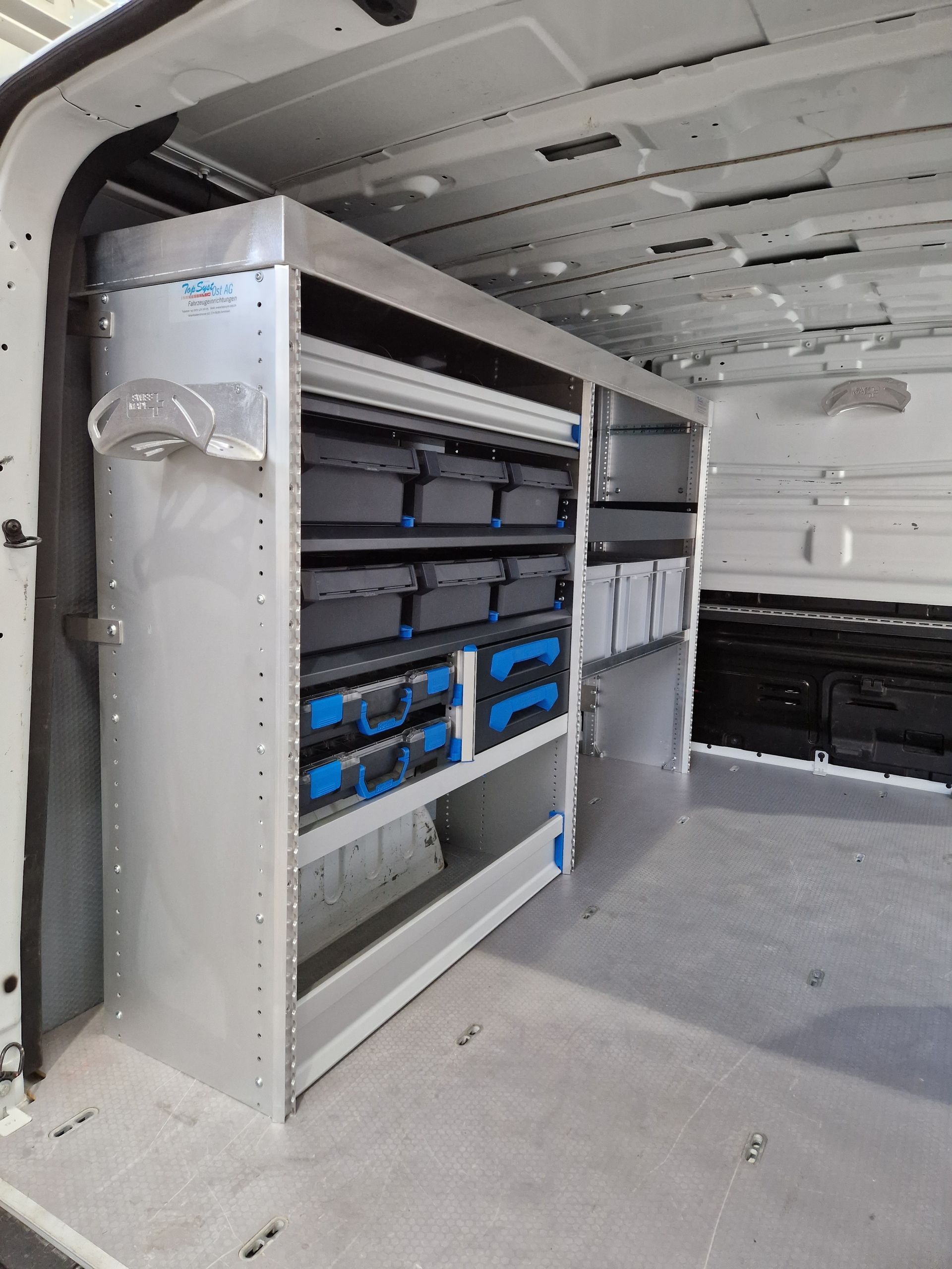 Innenraum eines weißen Lieferwagens mit Metallregalen für Werkzeuge und Lagerbehälter.