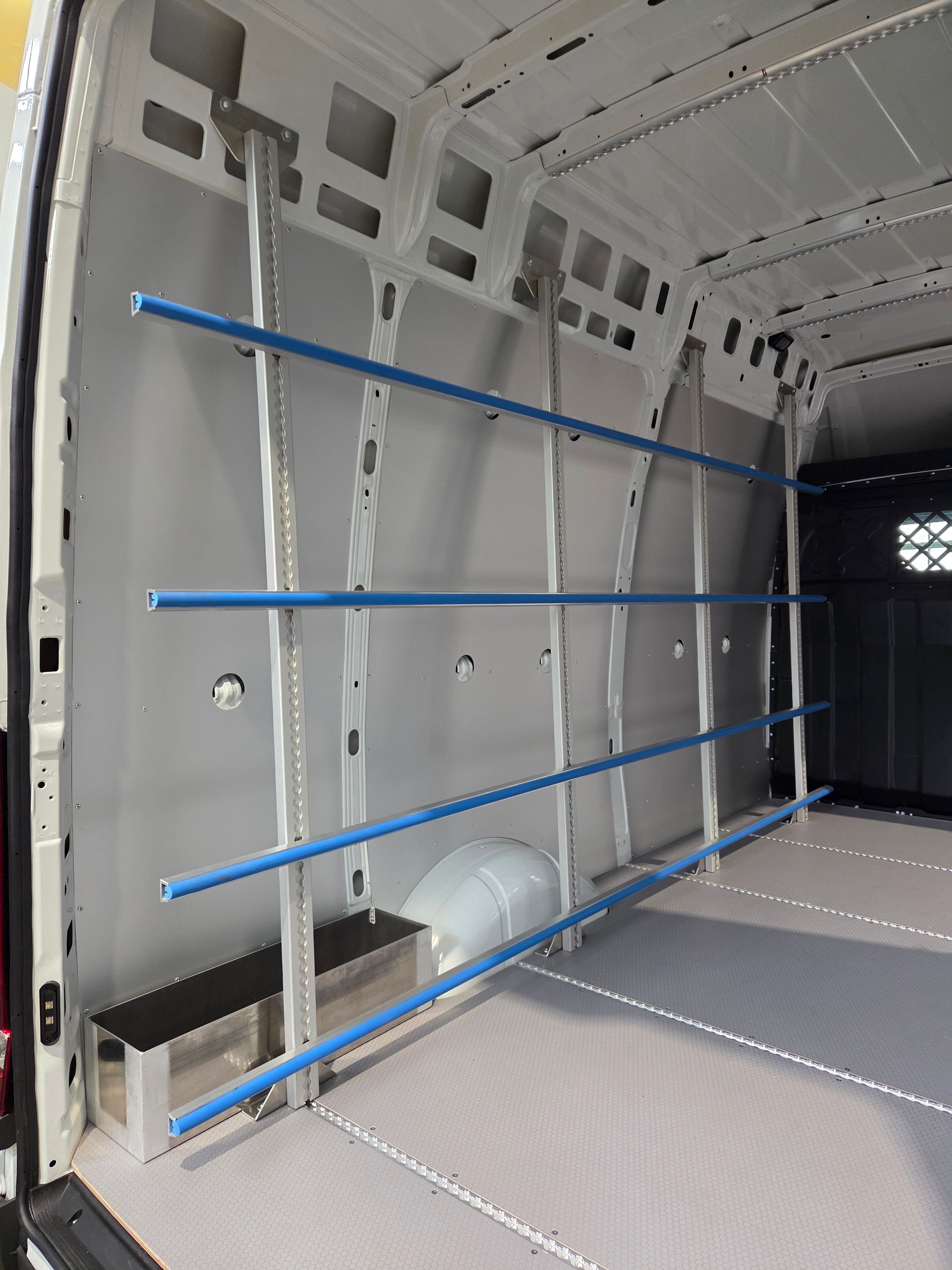 Innenraum eines weißen Lieferwagens mit Metallregalen und blauen Stützen, unten steht eine kleine Bank.